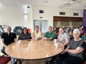 Trivallis Housing Landlord Wales Six people sit around a circular wooden table with white cups in front of them, smiling at the camera. Other people sit at tables in the background in a large, well-lit room.
