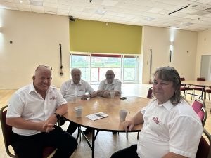 Trivallis Housing Landlord Wales Four men sit around a round table in a spacious room with light-colored walls and large windows. They are wearing white shirts and have cups and papers on the table in front of them.