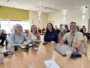 Trivallis Housing Landlord Wales Four adults, three women and one man, sit at a wooden table in a brightly lit room. The table has drinks, a laptop, notepad, and phones on it. Other people are visible in the background.