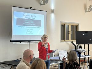 Trivallis Housing Landlord Wales A woman stands speaking in front of a seated audience in a room. Behind her is a screen displaying details for the “Tonyrefail FUND Day Celebration Event” at Tonyrefail Leisure Centre, along with a projector and sound equipment.