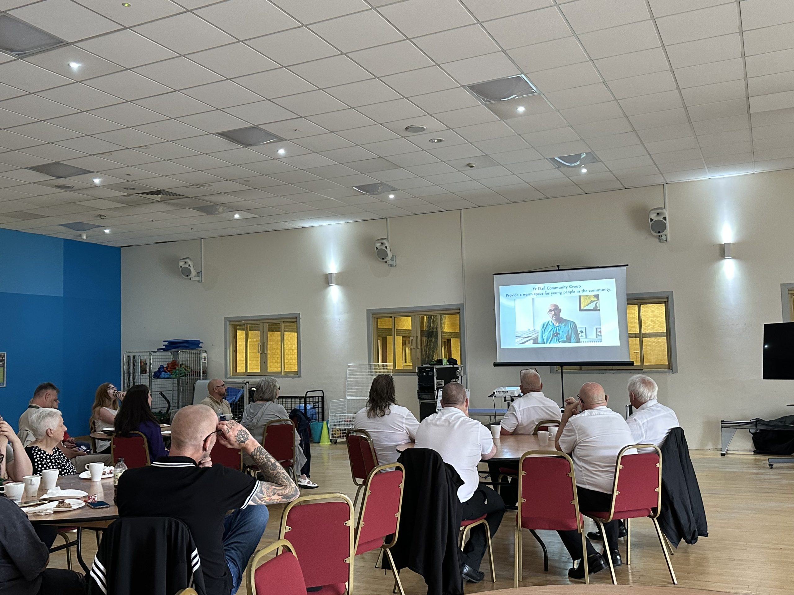 Trivallis Housing Landlord Wales A group of people sit on red chairs in a hall, facing a projector screen showing a presentation. Some attendees have drinks on the tables, and the room has a high ceiling with white walls and windows.