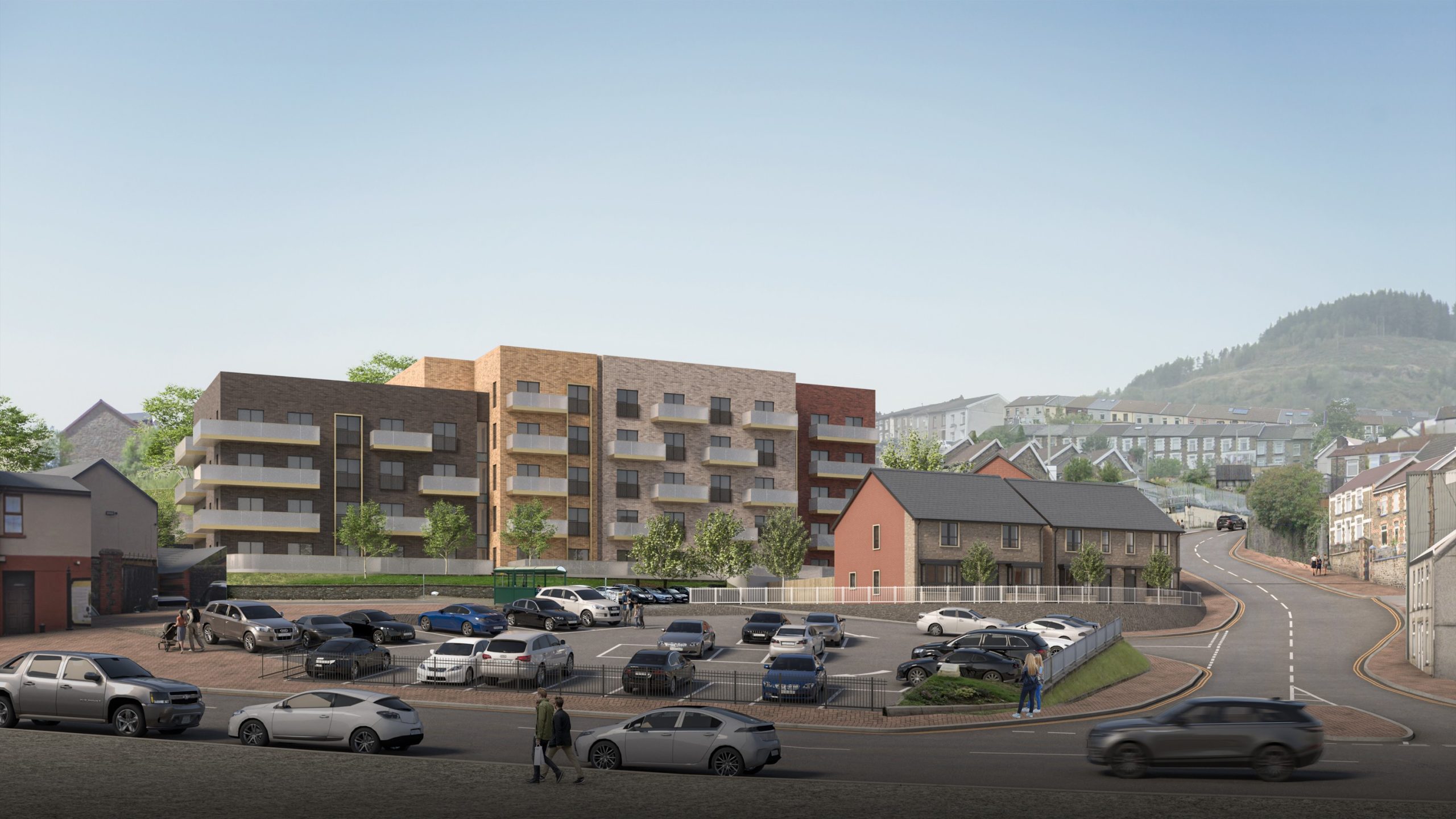 Trivallis Housing Landlord Wales A modern apartment building with balconies is situated behind a parking lot filled with cars, next to smaller houses; people walk along the street, and hills are visible in the background.