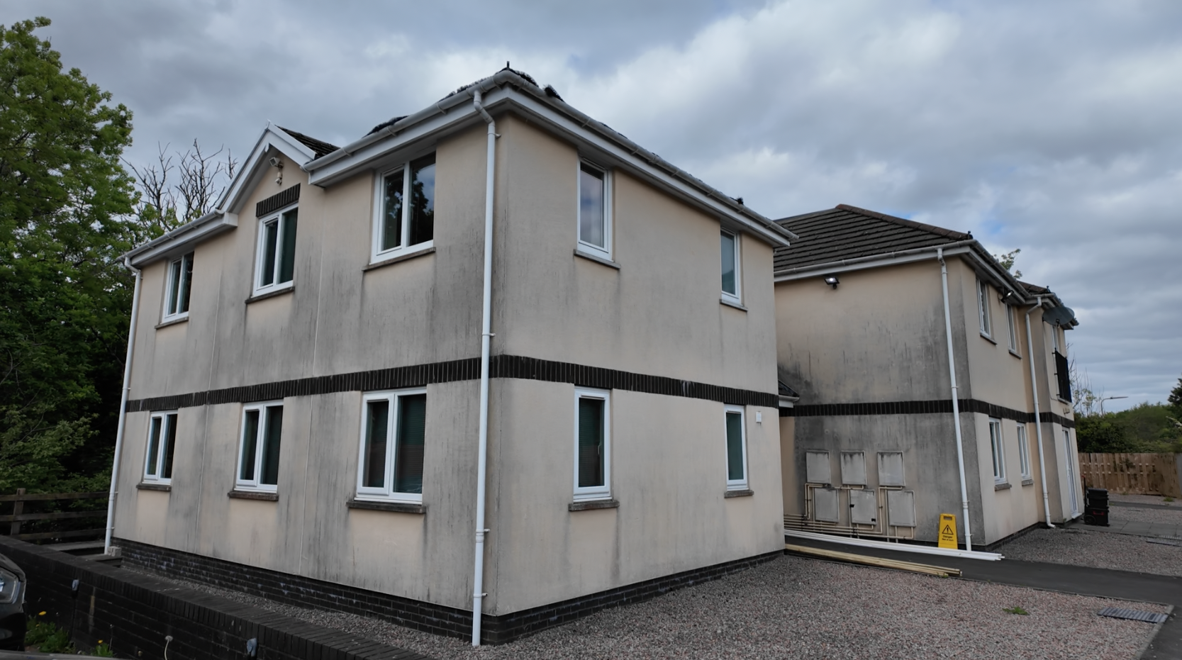 Trivallis Housing Landlord Wales Two modern, two-story cream-colored apartment buildings with white-trimmed windows. The building on the right has visible black staining on the walls. A cloudy sky is overhead, and there are some trees and a gravel yard.