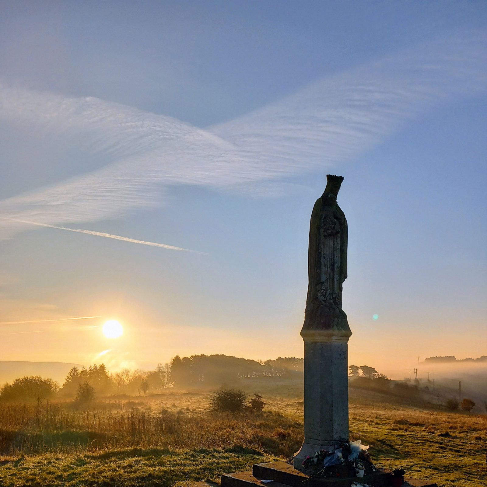 Trivallis Housing Landlord Wales A stone statue on a pedestal stands in a grassy field at sunrise, with flowers at its base. The sky is clear with scattered clouds, and mist covers the distant landscape and trees.