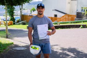 Trivallis Housing Landlord Wales A man wearing a grey t-shirt and shorts stands outdoors holding a white hard hat in one hand and a certificate in the other. There are houses, a fence, and trees in the background.