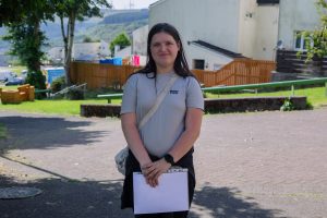 Trivallis Housing Landlord Wales A person with long brown hair stands outdoors holding a folder of papers. They wear a light grey t-shirt, black pants, and a watch, with houses, a fence, and greenery visible in the background.