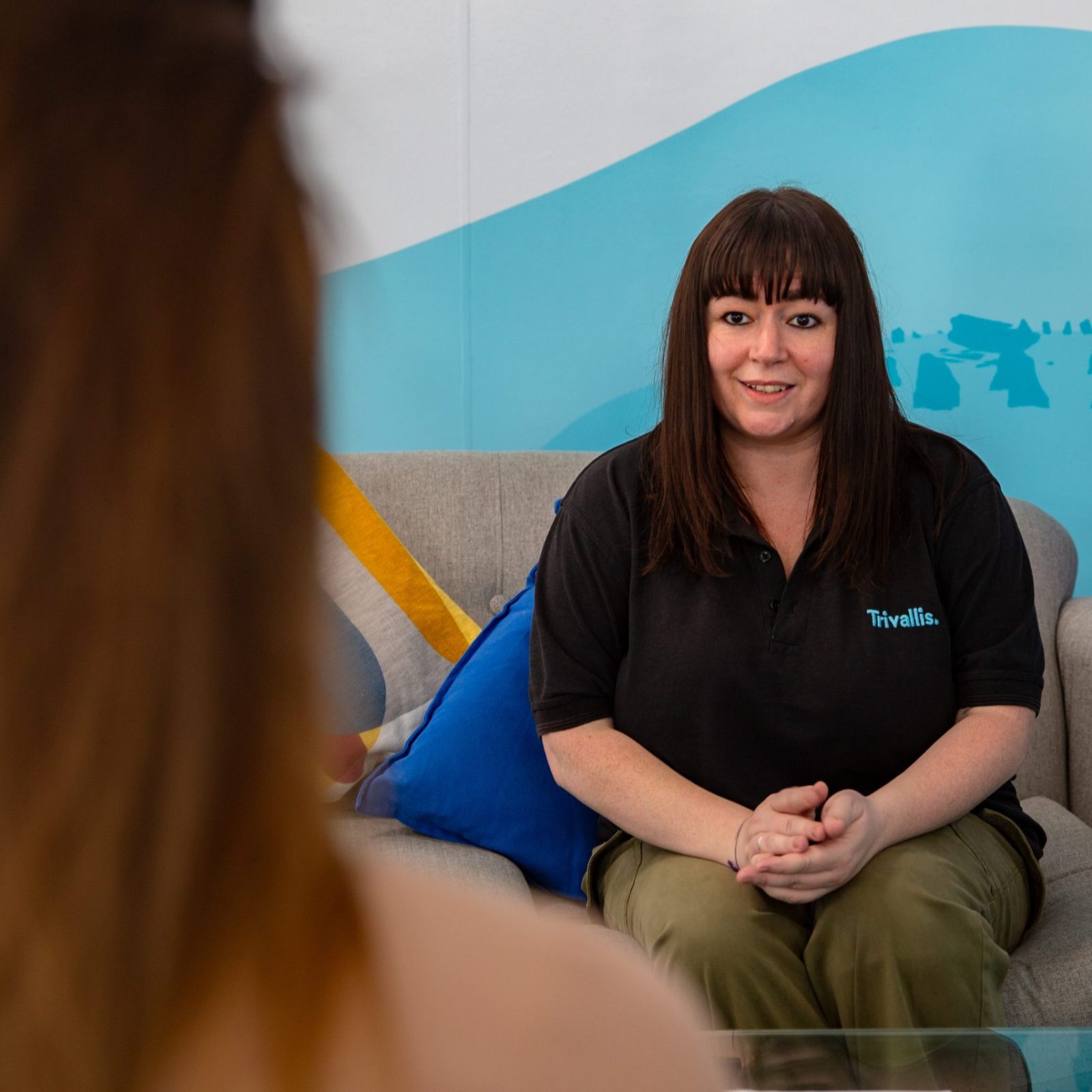 Trivallis Housing Landlord Wales A woman with straight brown hair and bangs, wearing a black shirt, sits on a gray couch and faces another person whose back is visible in the foreground. Colorful banners are visible in the background.