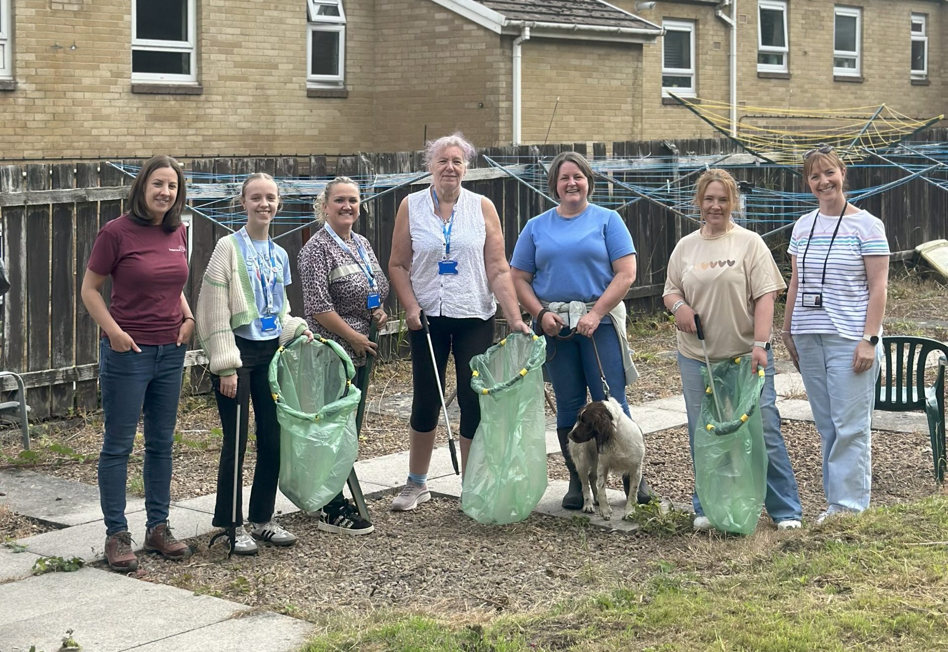 Trivallis Housing Landlord Wales Seven women stand outdoors in front of a building, holding green litter pickers and bags. A small dog is at their feet. The scene is overcast, and they appear to be participating in a cleanup activity.