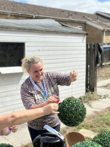 Trivallis Housing Landlord Wales A woman with light hair tied back is holding up a round artificial topiary ball outdoors near a white shed. Another person’s hand is visible assisting her. The scene takes place on a sunny day.