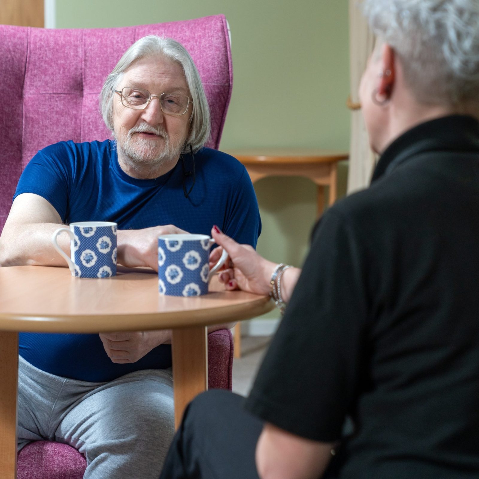 Trivallis Housing Landlord Wales An older man with long gray hair and glasses sits in a pink chair at a small wooden table with a woman. They both have mugs in front of them and appear to be having a conversation indoors.