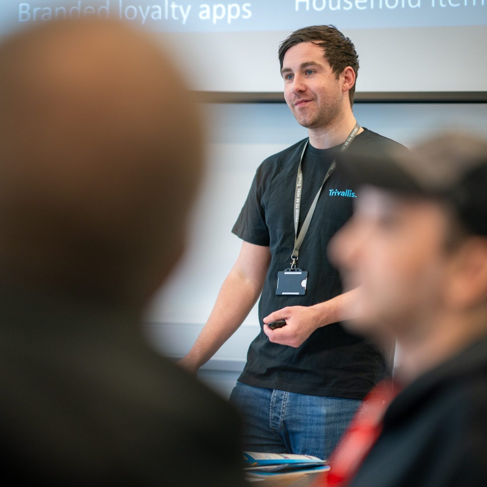 Trivallis Housing Landlord Wales A man in a black t-shirt stands at the front of a room, speaking to two seated people. A presentation is projected on a screen behind him, displaying partially readable text about apps and loyalty programs.