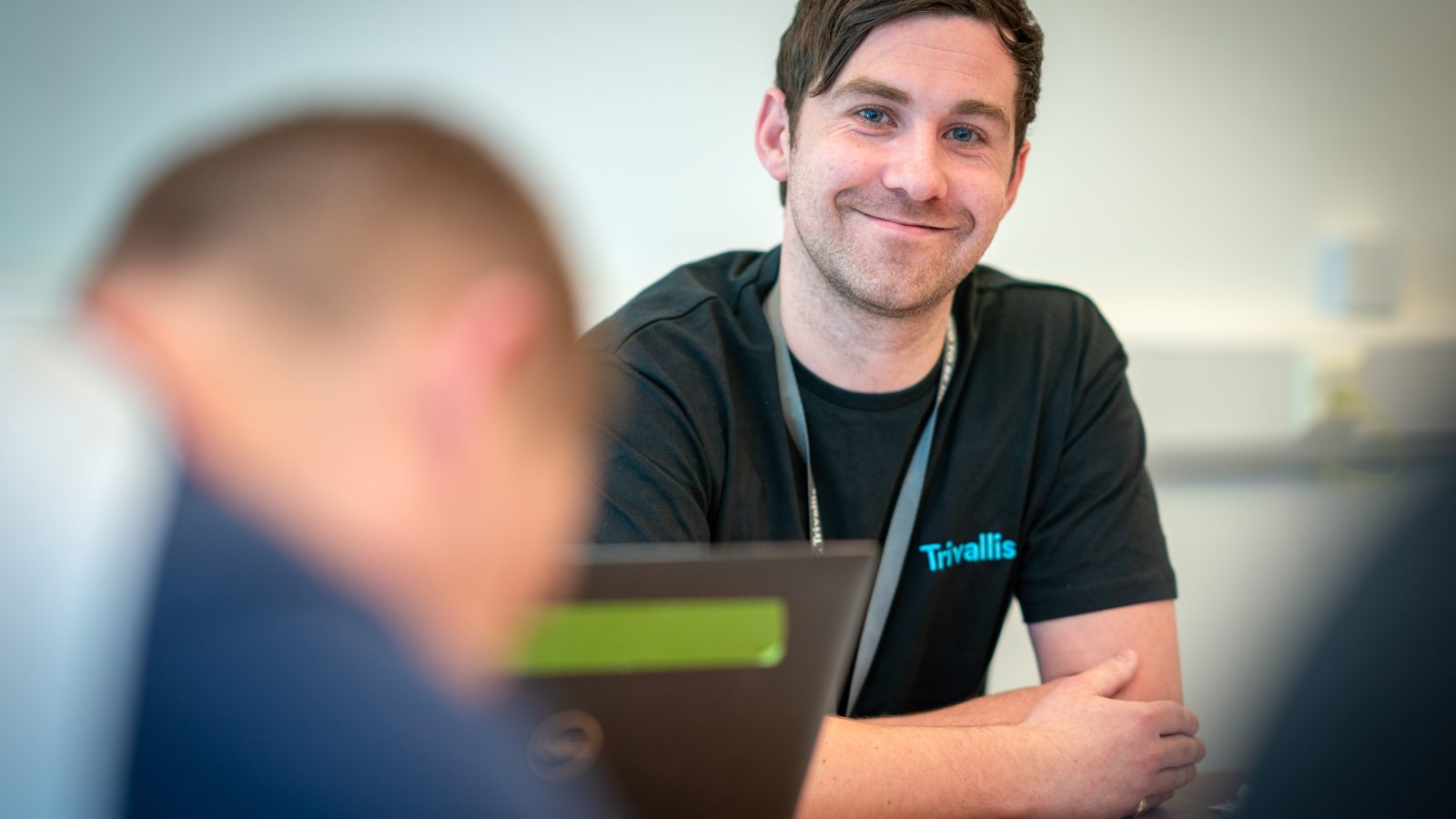 Trivallis Housing Landlord Wales A man wearing a black Trivallis t-shirt sits at a desk smiling at the camera, with a laptop and papers in front of him. Another person is blurred in the foreground.