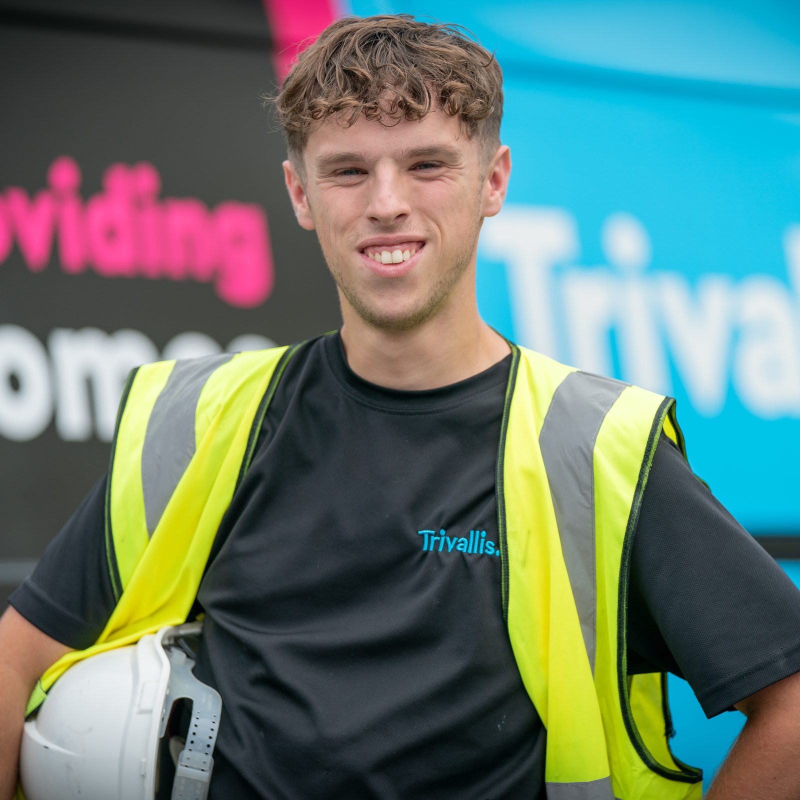 Trivallis Housing Landlord Wales A young man wearing a yellow safety vest and holding a white hard hat stands in front of a van with the Trivallis logo and partial text 