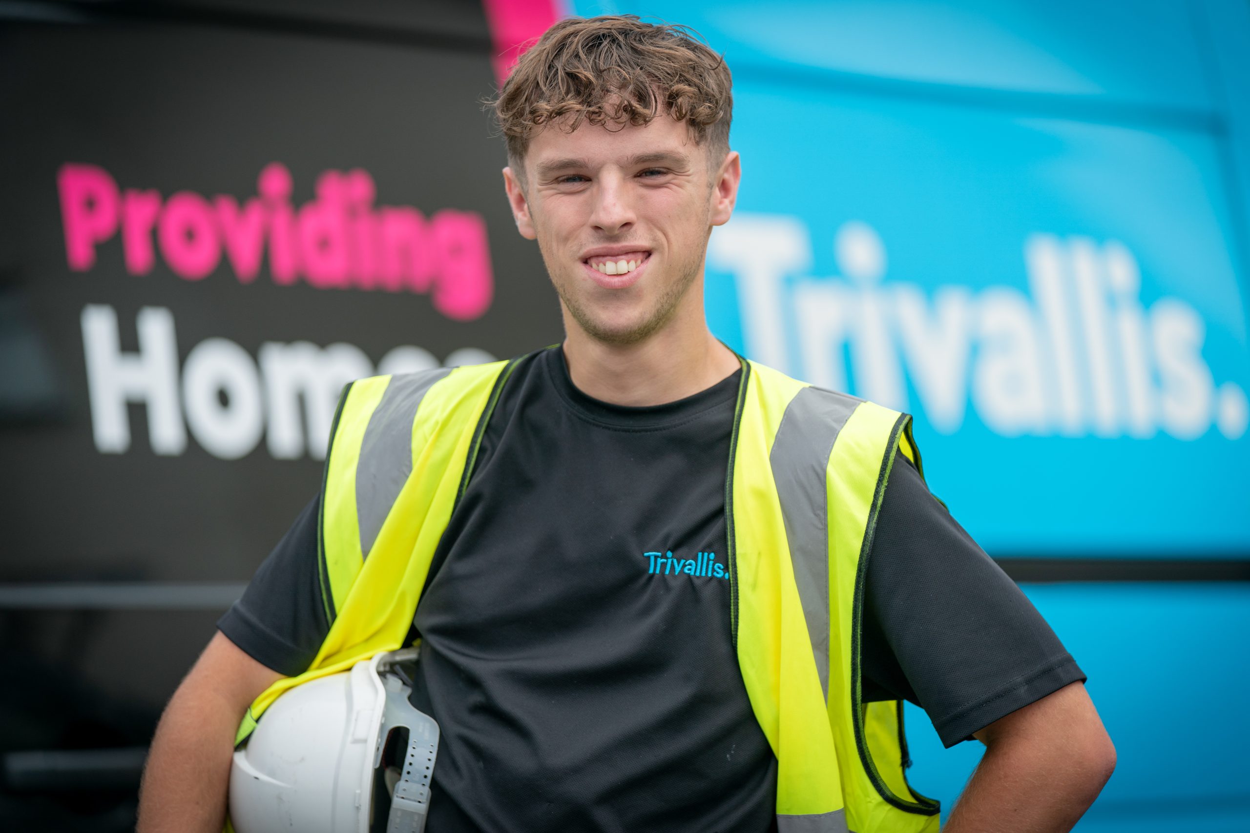 Trivallis Housing Landlord Wales A young man wearing a yellow safety vest and holding a white hard hat stands in front of a van with the Trivallis logo and partial text "Providing Homes" visible on its side.
