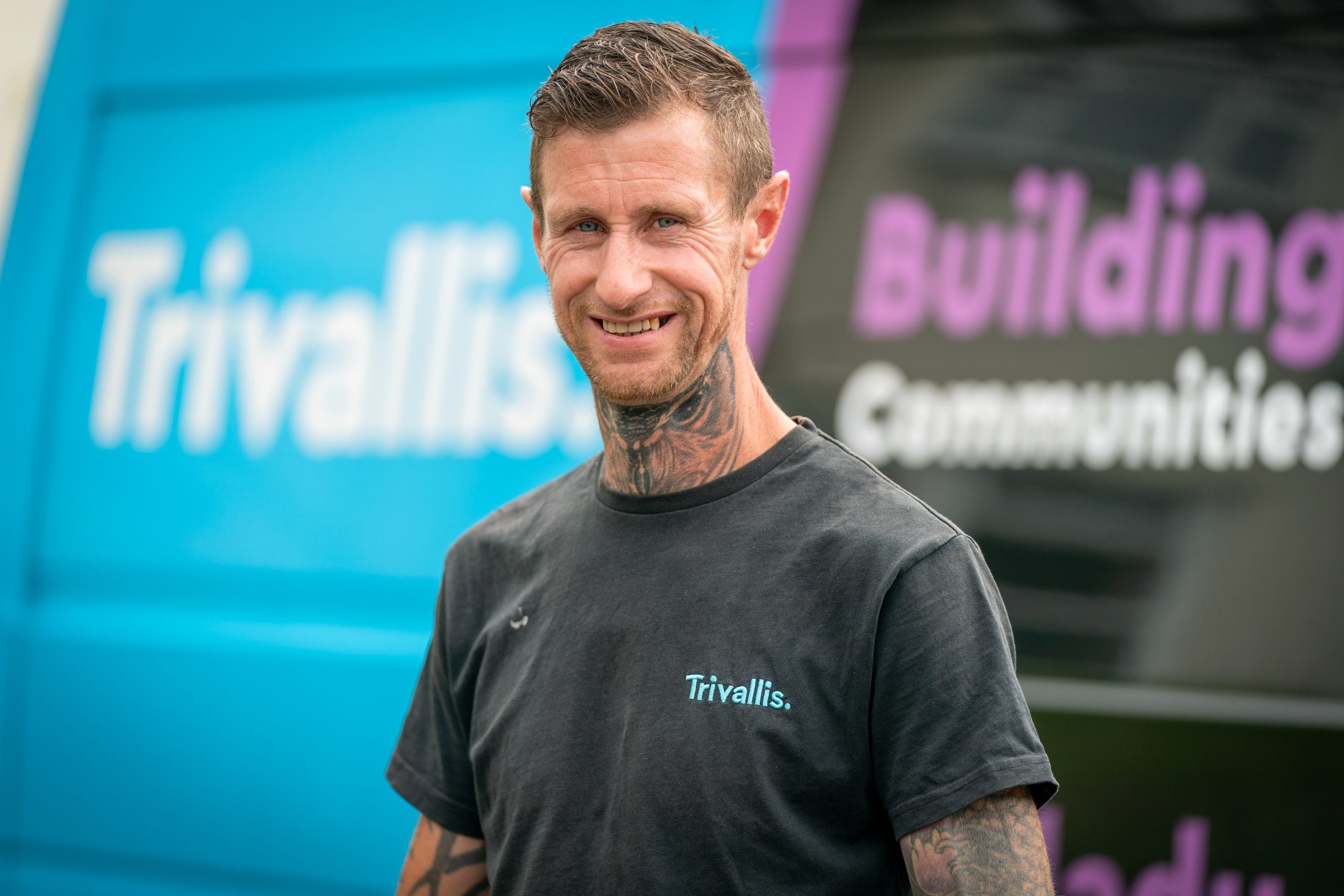 Trivallis Housing Landlord Wales A man with tattoos on his neck and arms wears a black "Trivallis" t-shirt and stands in front of a blue and purple Trivallis van. He is smiling at the camera.