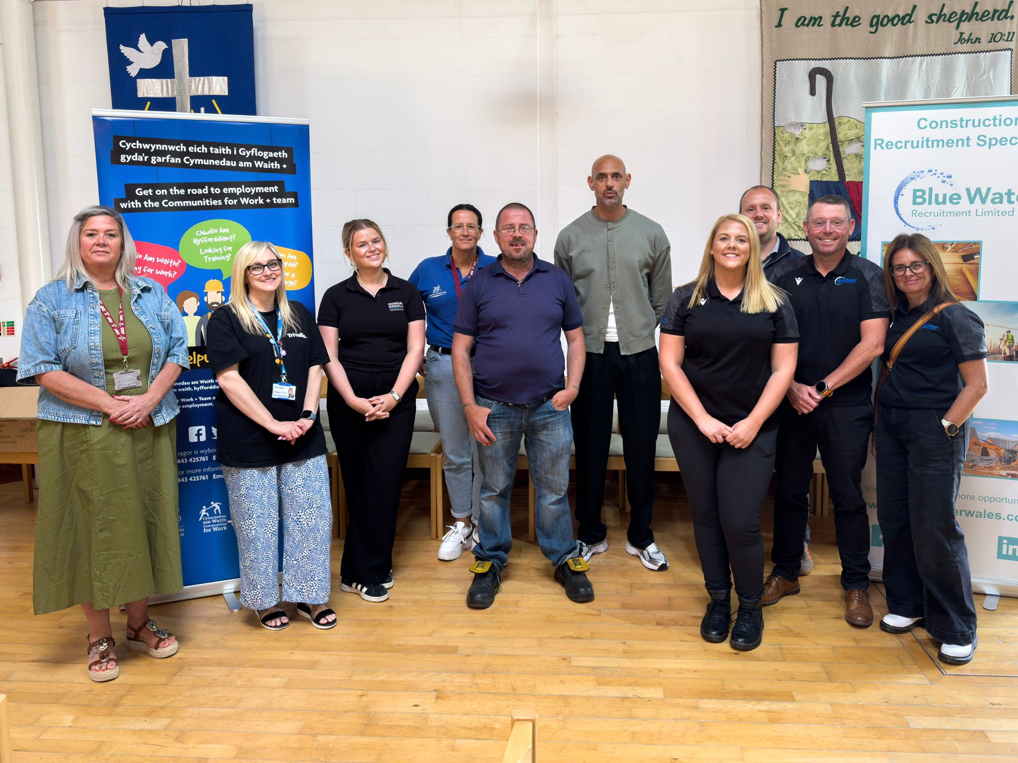 Trivallis Housing Landlord Wales A group of ten people stand indoors on a wooden floor, facing the camera. They are positioned between two informational banners and appear to be at an event or seminar. The background includes a sign and part of a table.