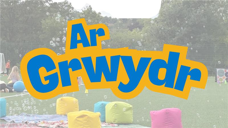 Trivallis Housing Landlord Wales Text "Ar Grwydr" in large, bold blue and yellow letters over a background showing an outdoor grassy area with children, colorful beanbags, and bubbles. Trees and hills are visible in the distance.