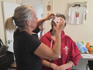 Trivallis Housing Landlord Wales A woman applies face paint to a girl wearing glasses and a red hoodie. Balloons, a coloring sheet, and a t-shirt with a fries graphic are visible in the background of a room.