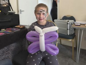 Trivallis Housing Landlord Wales A young child with face paint sits indoors, smiling and holding a large balloon shaped like a butterfly with white and purple sections. She is wearing a grey outfit with white polka dots.