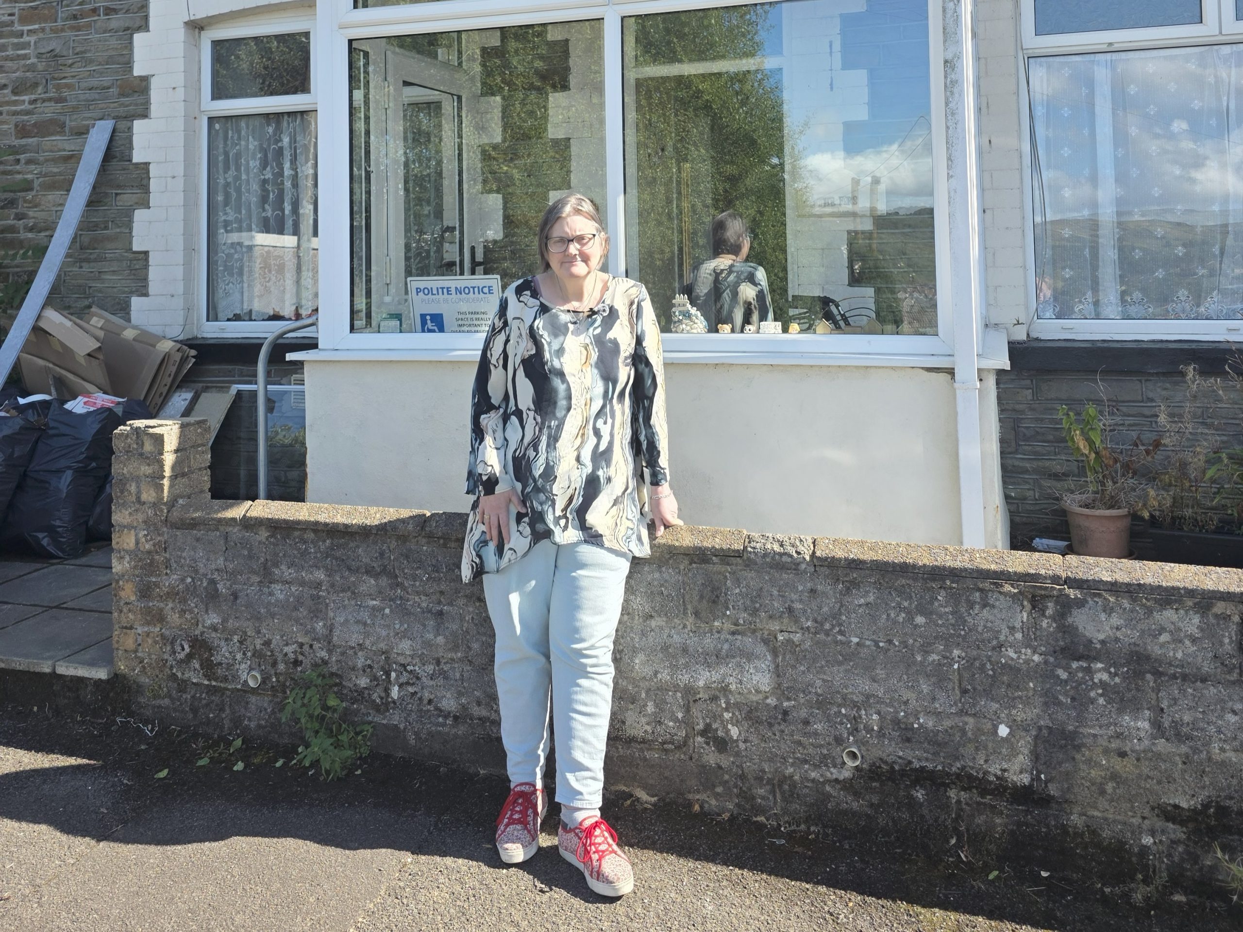 Trivallis Housing Landlord Wales A person with light-colored hair, glasses, a patterned top, light jeans, and red shoes stands in front of a low stone wall and a house with large windows. There are bags and items stacked on the left side.