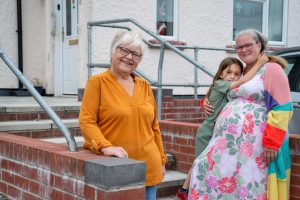 Trivallis Housing Landlord Wales Three people stand outside a house. An older woman in an orange shirt stands by a brick wall, while a woman in a floral dress and colorful cardigan hugs a child. Steps and a handrail lead up to the front door behind them.