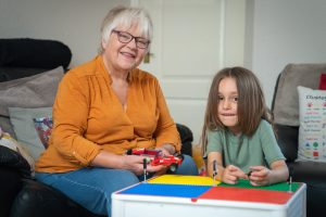 Trivallis Housing Landlord Wales An older woman and a young child sit together indoors, smiling and playing with toy cars on a large, colorful building block base. Cushions and a door are visible in the background.