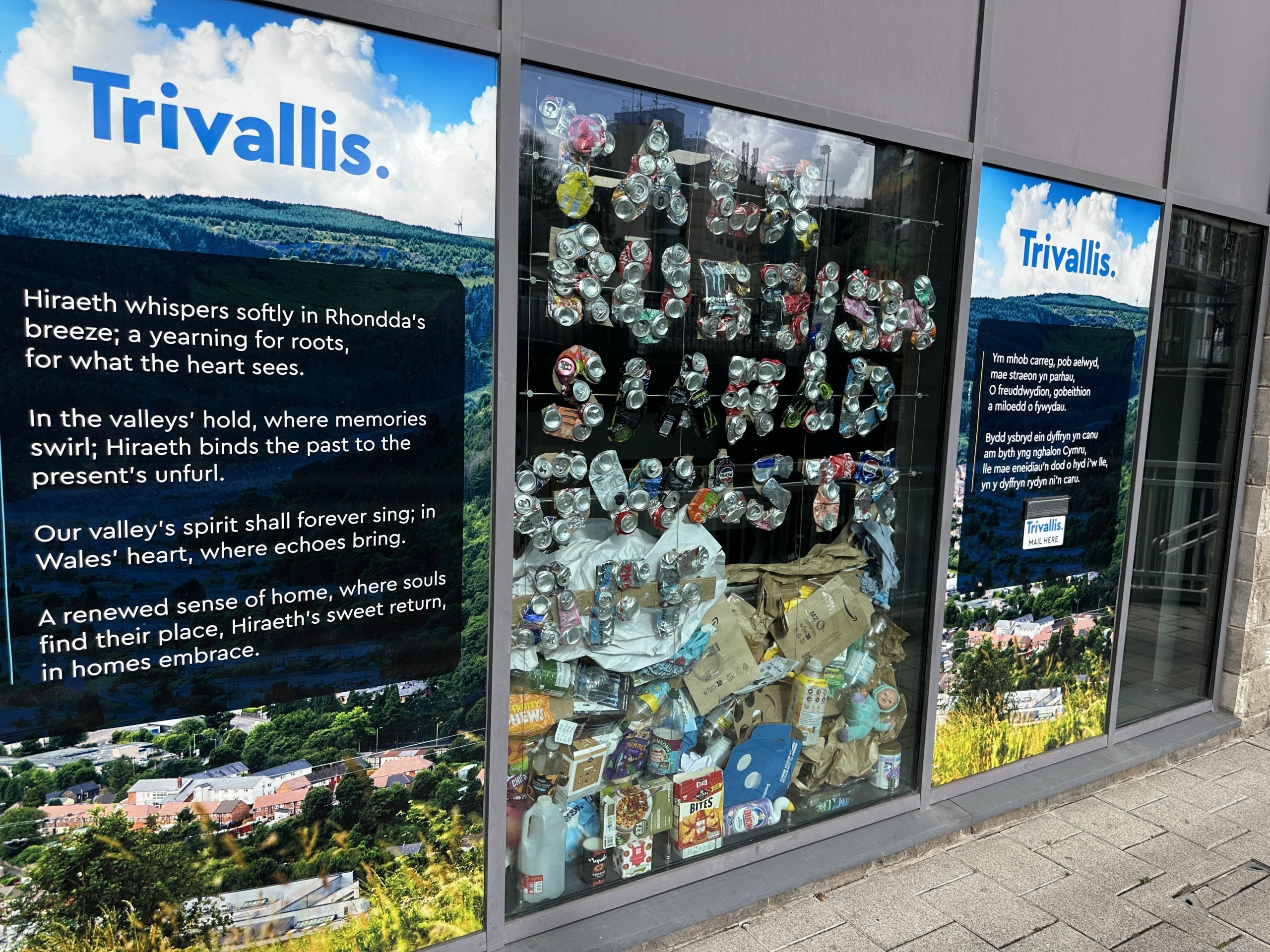 Trivallis Housing Landlord Wales A window display features a large pile of crushed cans and plastic bottles. Surrounding windows display blue posters with the Tryalls logo and white text about the environment. The image is rotated sideways.