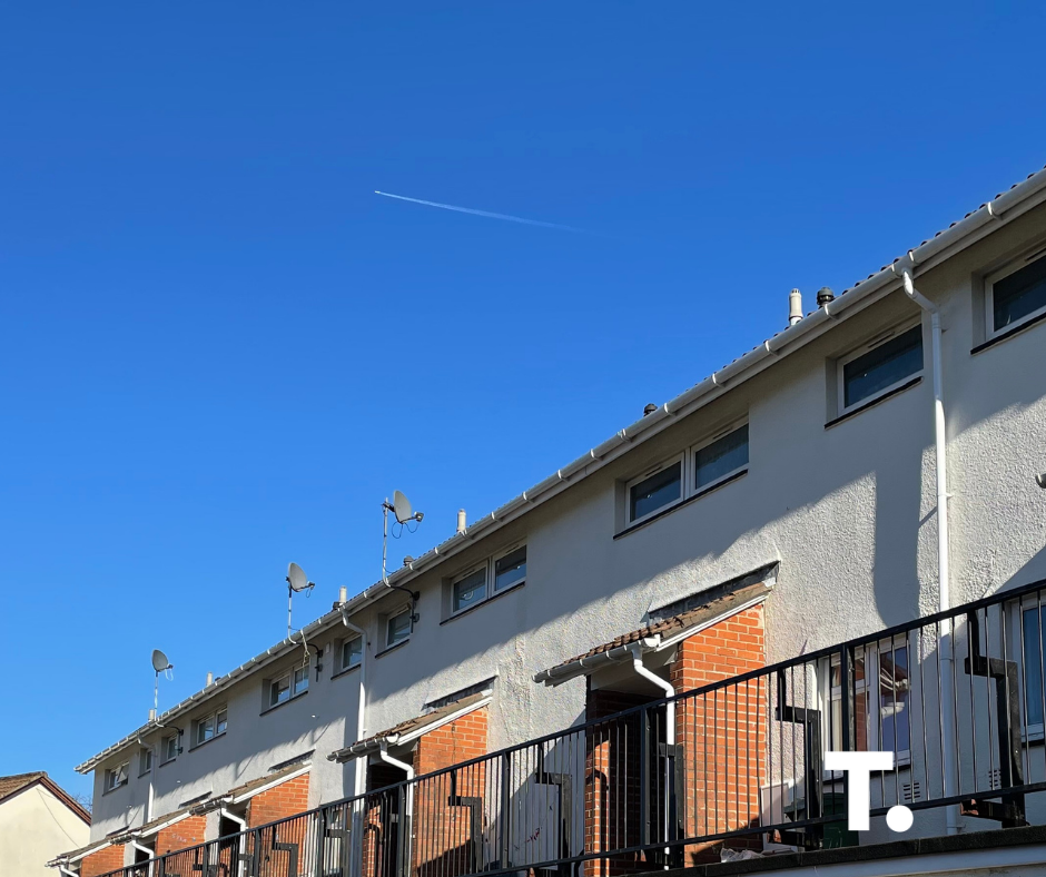 Trivallis Housing Landlord Wales A row of modern townhouses with white and brick exteriors, rooftop satellite dishes, and black railings. The sky above is clear and blue, with a visible airplane contrail. A white "T." logo is in the lower right corner.