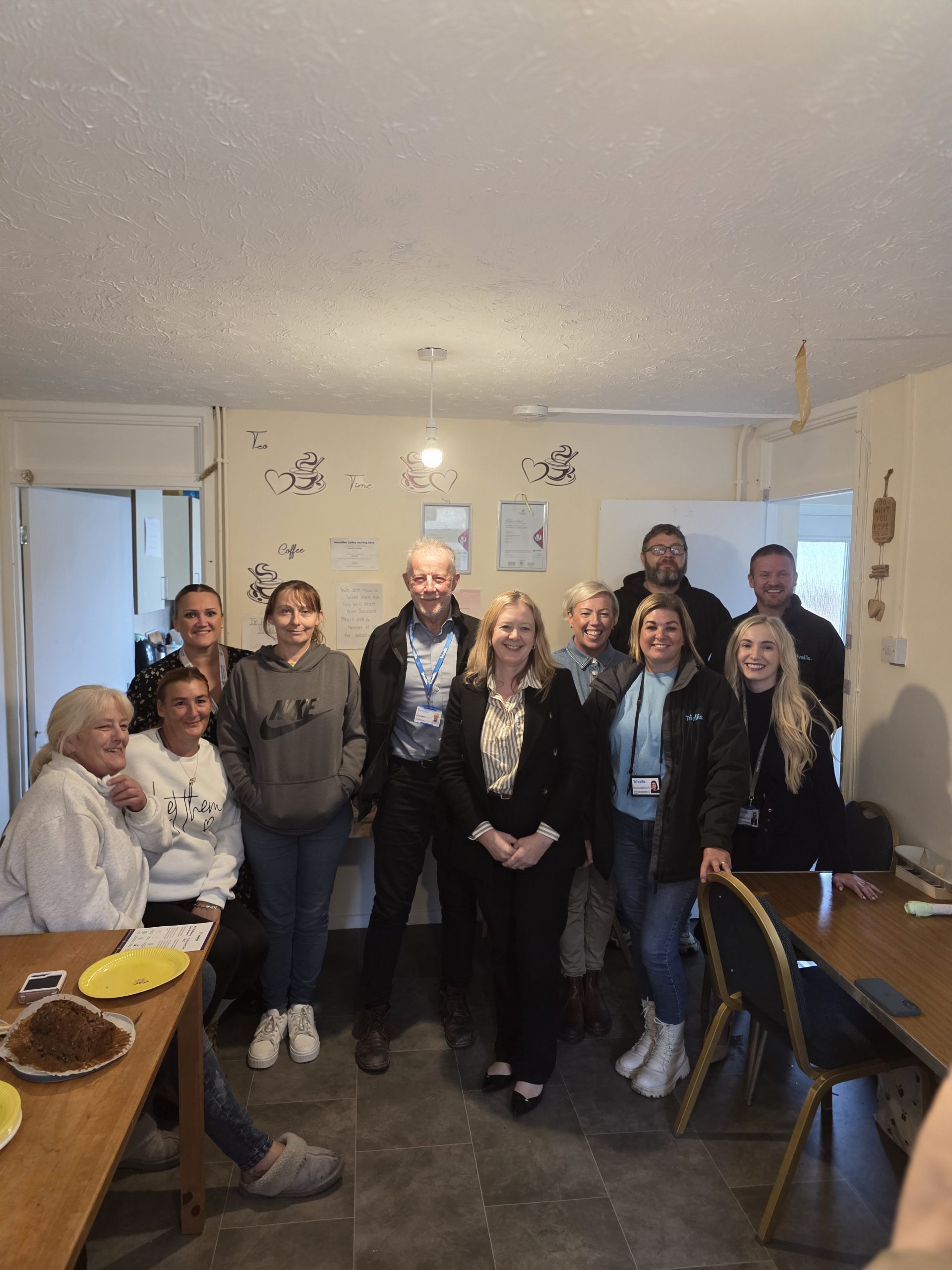 Trivallis Housing Landlord Wales A group of eleven people pose together in a kitchen or dining area, some standing and some sitting. There are plates and a cake on the table, and handwritten notes or drawings are visible on the wall behind them.