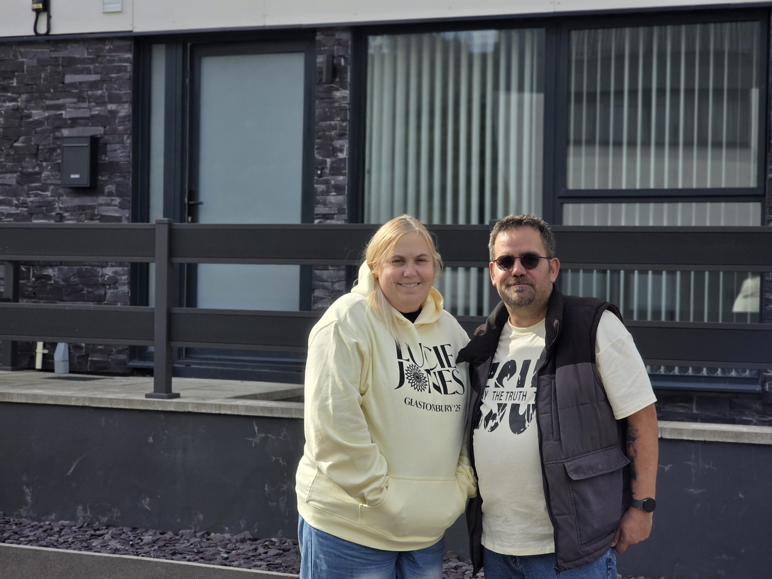 Trivallis Housing Landlord Wales Two people stand close together outside a modern building with gray stone and large windows. The person on the left wears a cream hoodie and jeans; the person on the right wears sunglasses, a t-shirt, and a vest.
