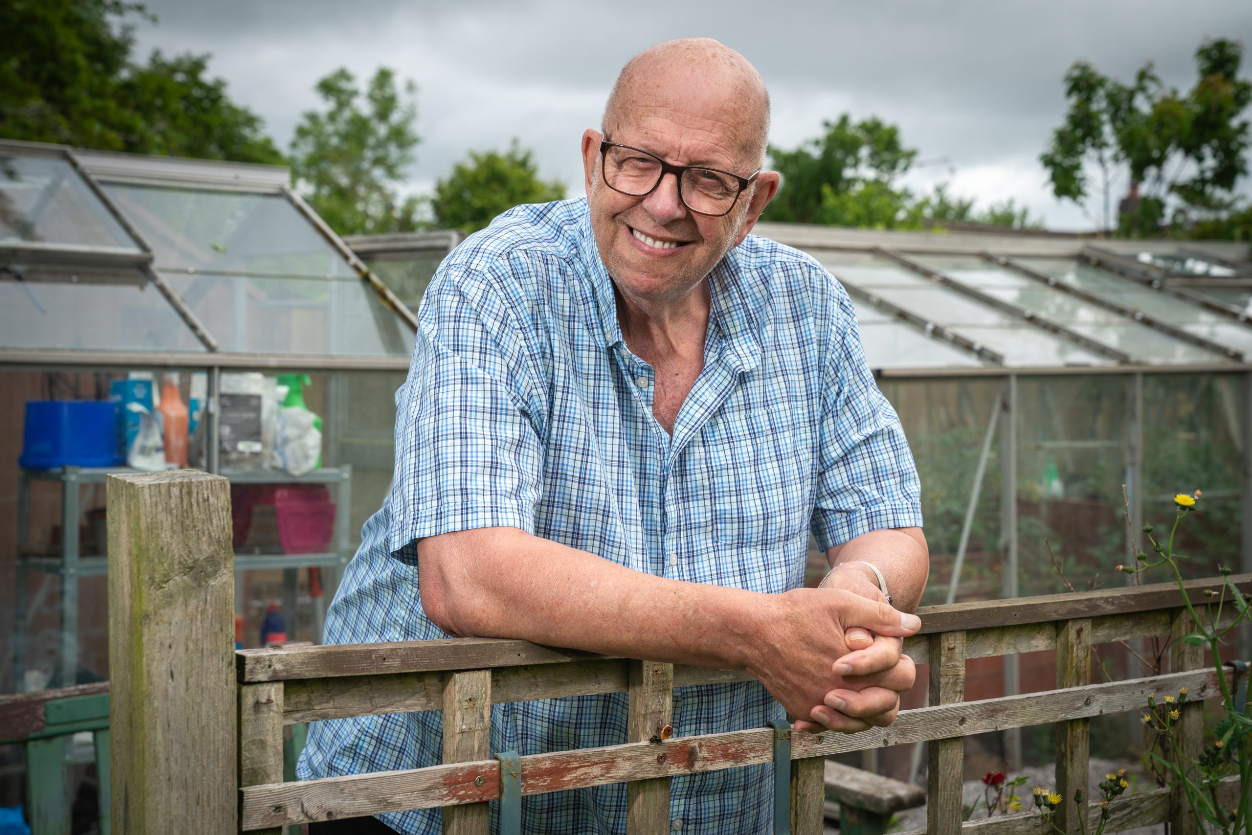 Trivallis Housing Landlord Wales An older man wearing glasses and a blue plaid shirt leans on a wooden fence, smiling, with greenhouses and plants in the background on a cloudy day.