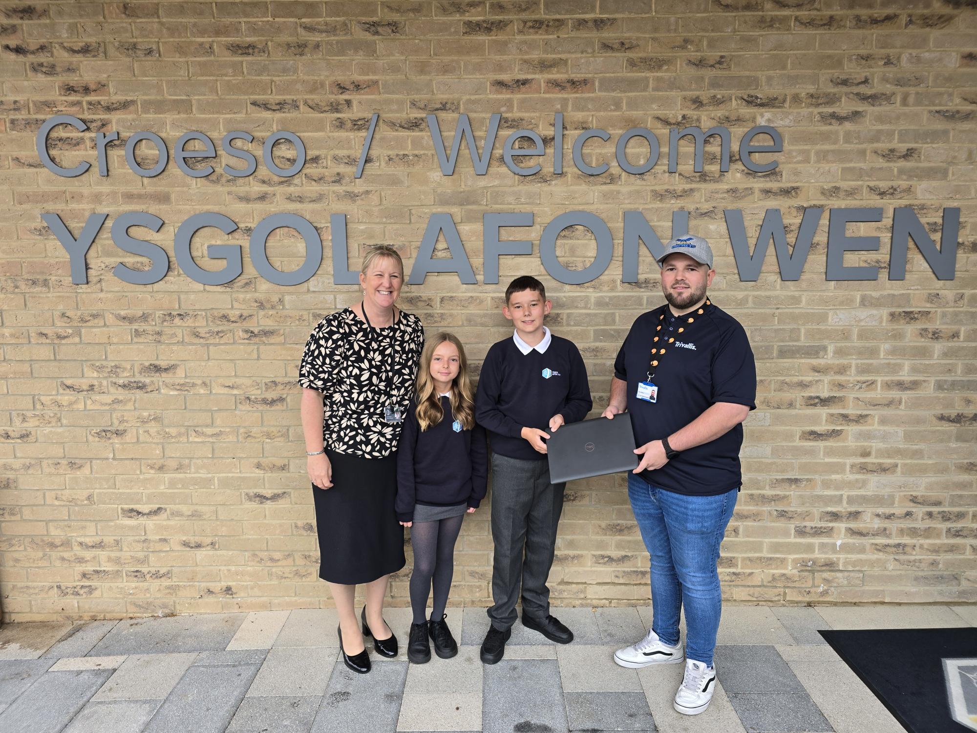 Trivallis Housing Landlord Wales Four people stand in front of a brick wall with the sign "Croeso / Welcome YSGOL AFON WEN." Two adults and two children are posed, with one adult handing a laptop to one of the children.