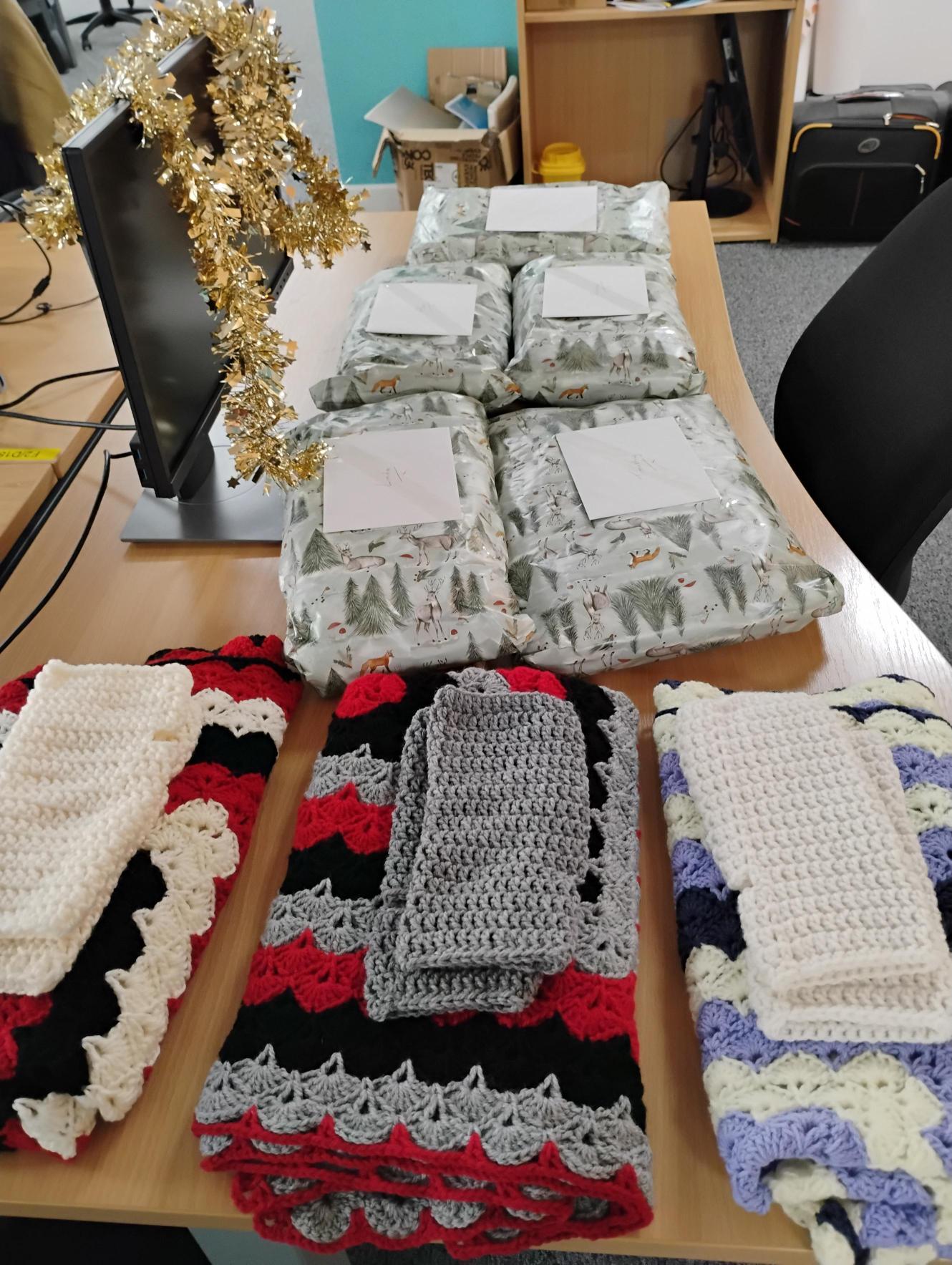 Trivallis Housing Landlord Wales Three crocheted blanket and towel sets are laid out on a table in the foreground, each with different color patterns. In the background, six wrapped packages labeled with cards are arranged near a computer decorated with tinsel.