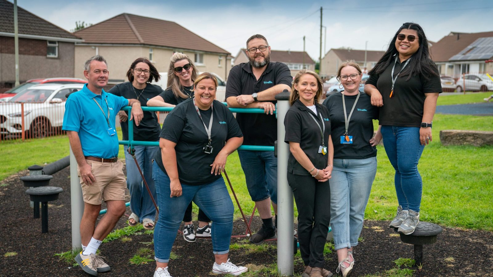 Trivallis Housing Landlord Wales Eight adults stand and smile at the camera near playground equipment in a residential area. Most are wearing matching black shirts and lanyards, while one man on the left wears a turquoise shirt. Houses and cars are in the background.