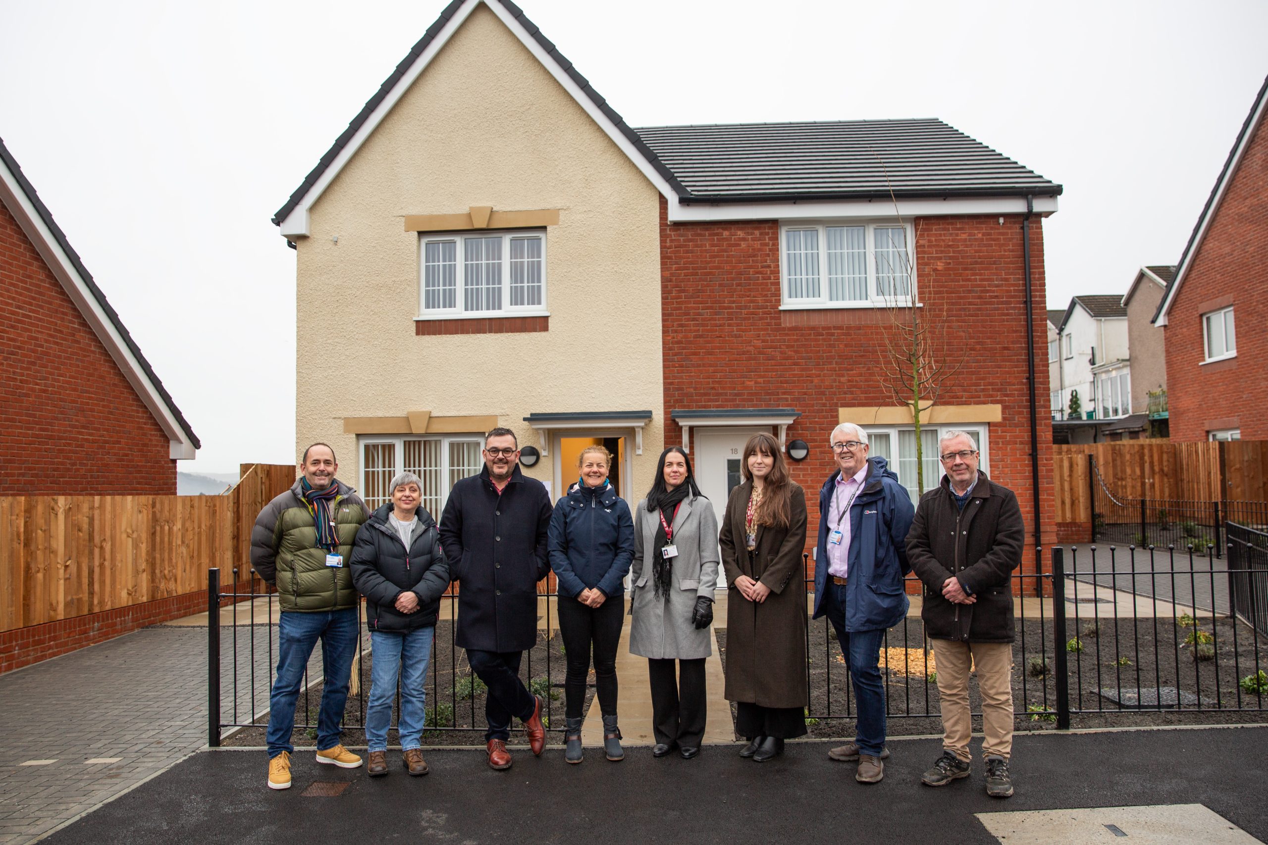 Trivallis Housing Landlord Wales Eight people stand in a row outside a modern two-story house with a cream and red brick exterior. The group is dressed in winter clothing and the weather appears overcast.