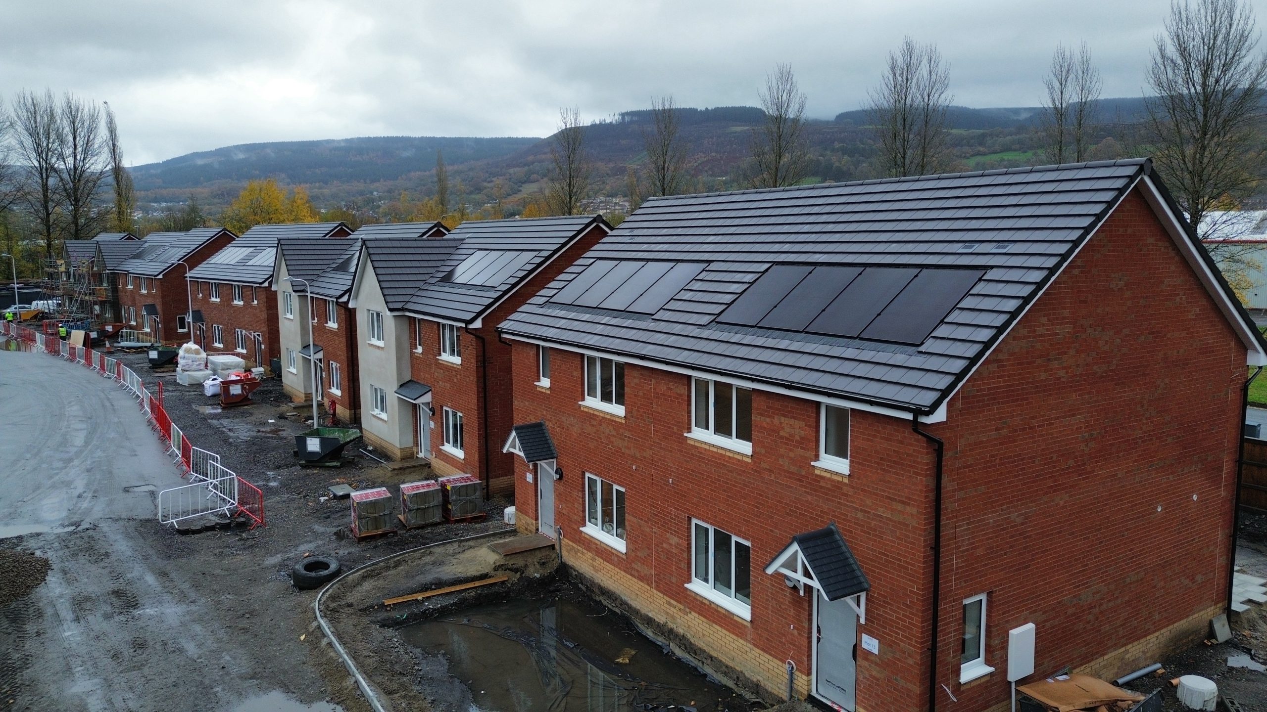 Trivallis Housing Landlord Wales A row of newly built brick houses with solar panels on the roofs, located on a muddy construction site with building materials and barriers, under a cloudy sky with hills in the background.