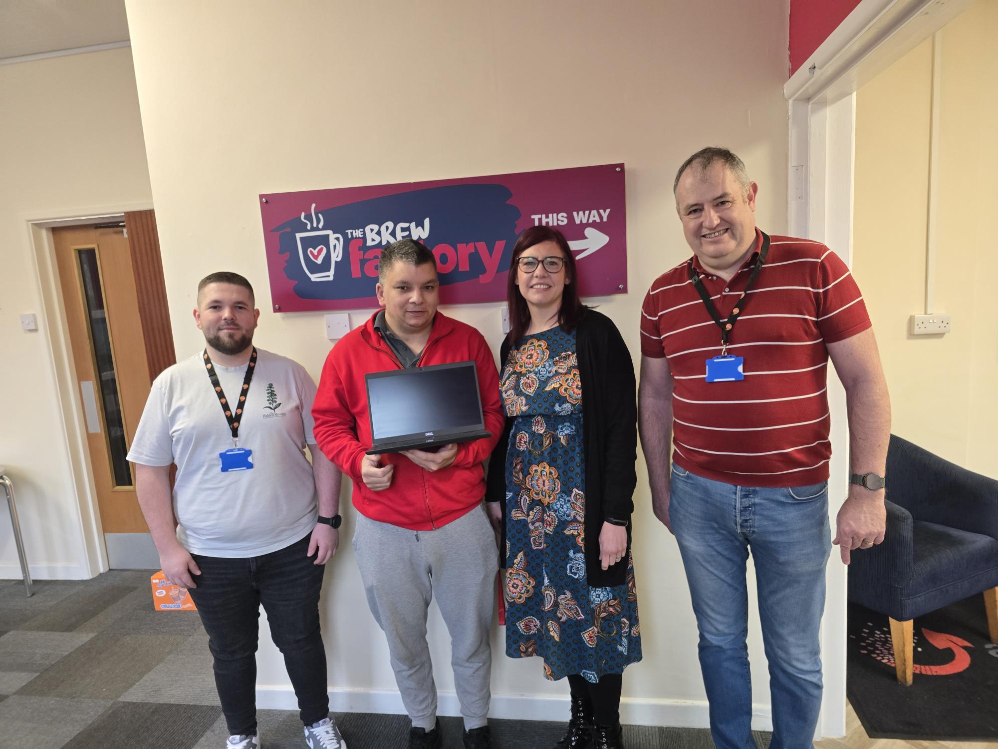 Trivallis Housing Landlord Wales Four people stand indoors in front of a sign that reads "The Brew Factory." One person, second from the left, holds a closed laptop. All four are wearing name badges and posing for the photo.