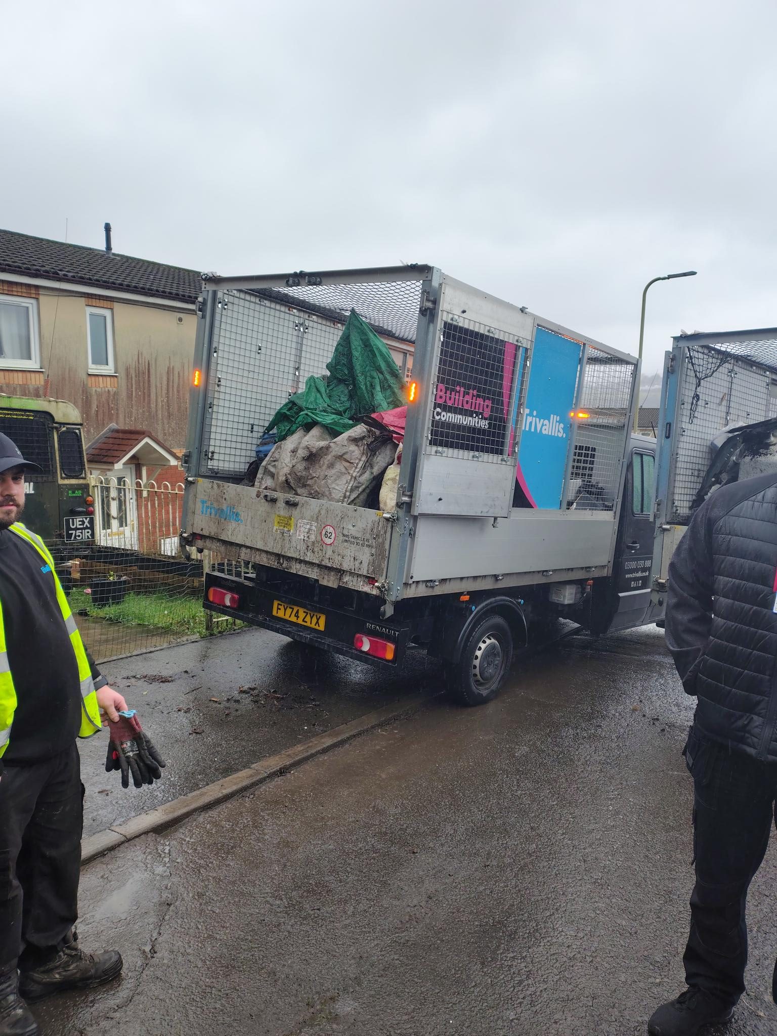 Trivallis Housing Landlord Wales A light truck with a Trivallis and Building Communities logo is parked on a wet residential street, carrying bags and debris. Two people in workwear stand nearby. The sky is overcast.