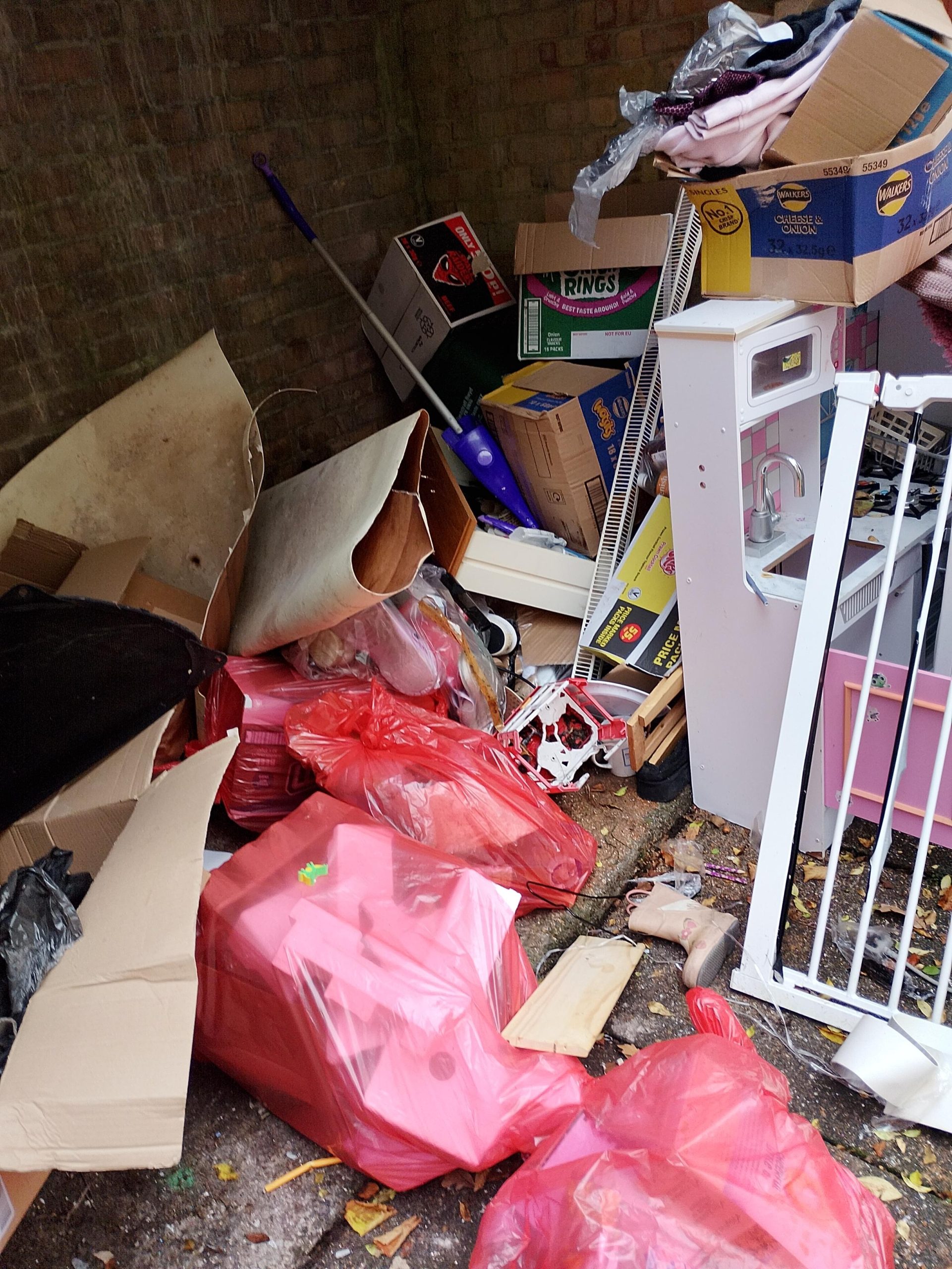 Trivallis Housing Landlord Wales A pile of discarded household items, cardboard boxes, red trash bags, and broken furniture is stacked haphazardly in a corner against a brick wall. The area appears cluttered and untidy.