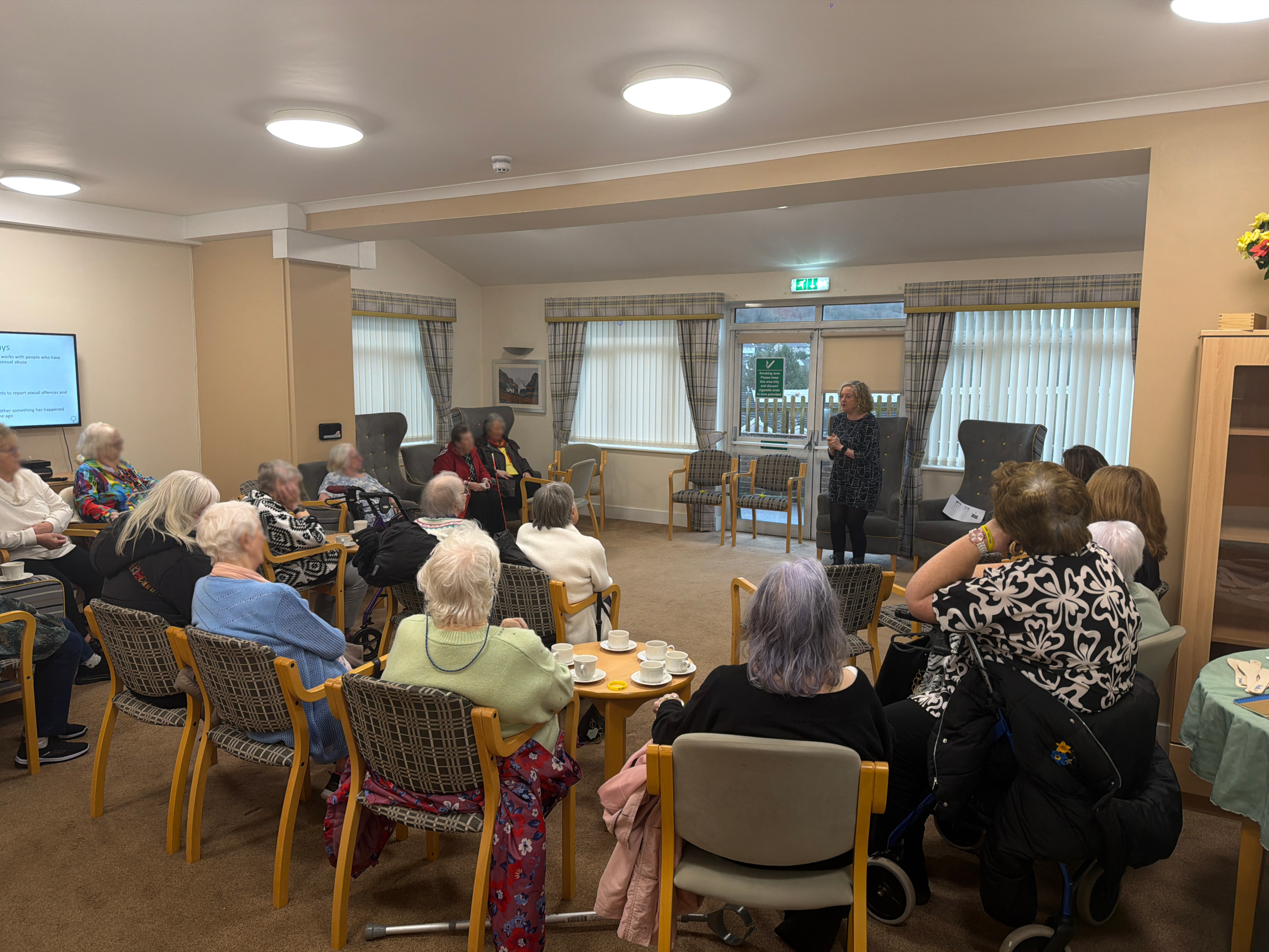 Trivallis Housing Landlord Wales A group of elderly people sit in a lounge, facing a woman standing and speaking at the front. Some people are in wheelchairs; tea cups and plates are on the tables. A few caregivers are present. Large windows are in the background.