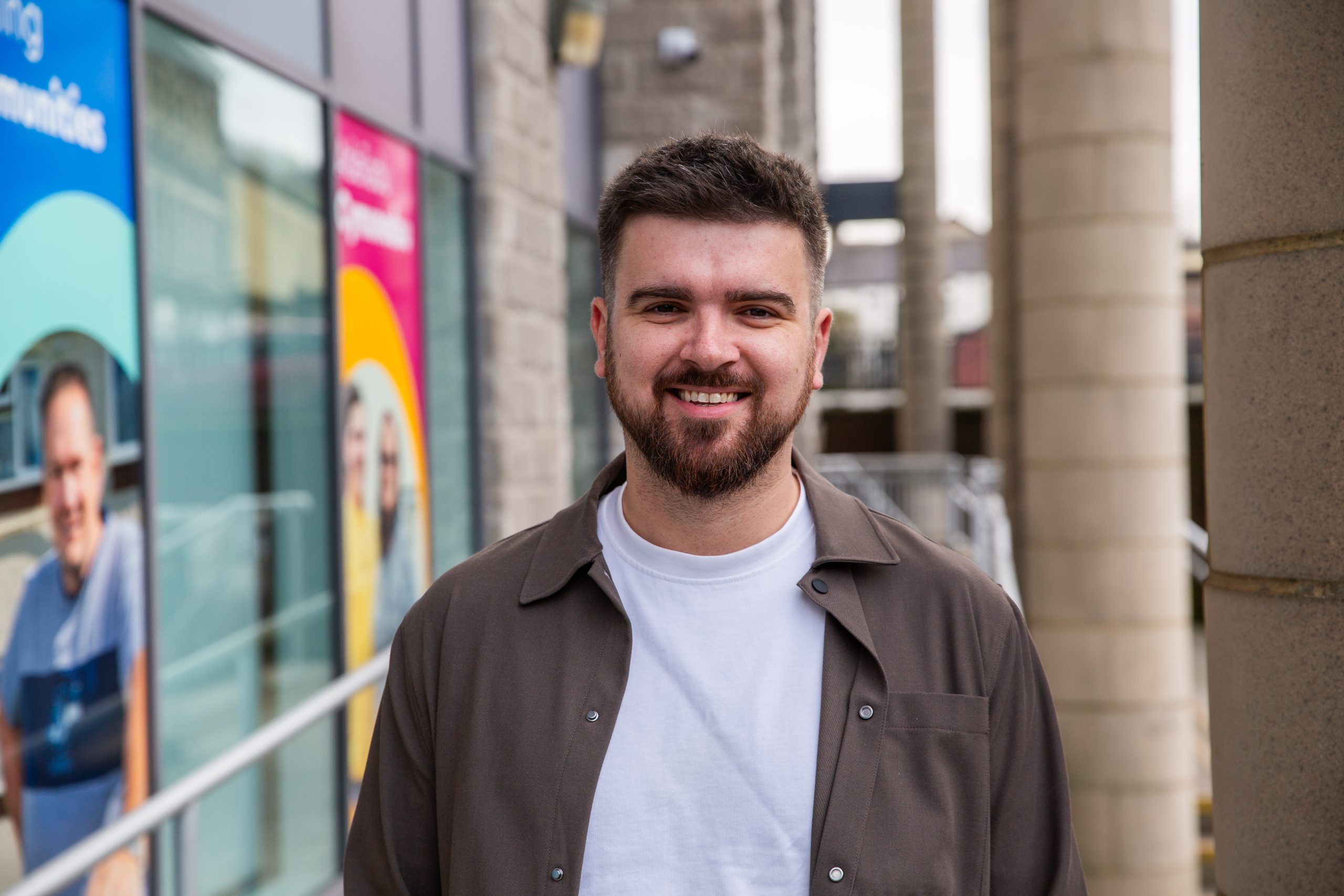 Trivallis Housing Landlord Wales A man with short dark hair and a beard is smiling at the camera. He is wearing a white t-shirt and an open brown shirt, standing outside near a building with colorful posters in the background.