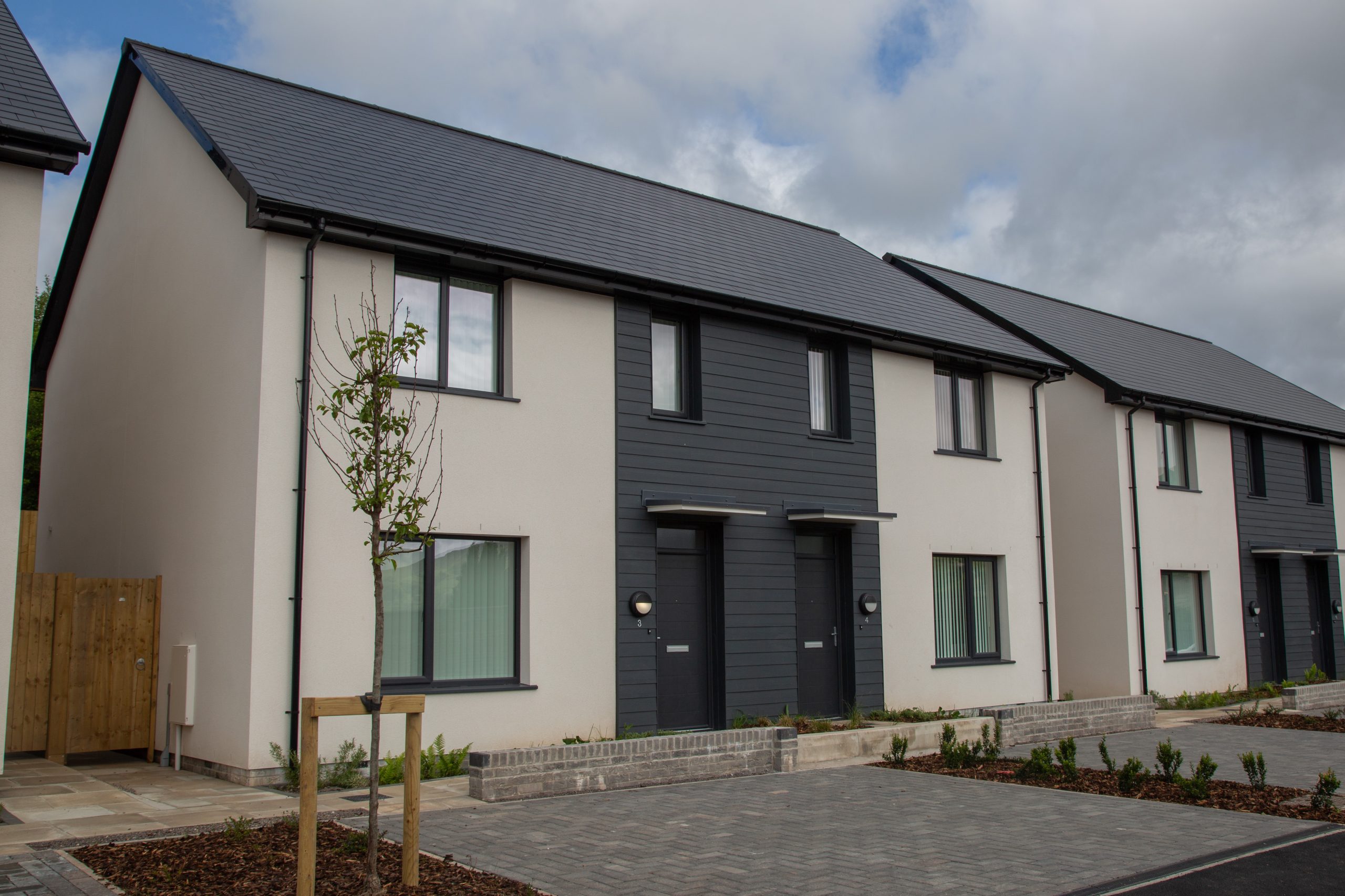 Trivallis Housing Landlord Wales A row of modern, semi-detached houses with light-colored walls, dark roofs, and small front gardens. The houses have large windows and individual entrances. The sky is partly cloudy.