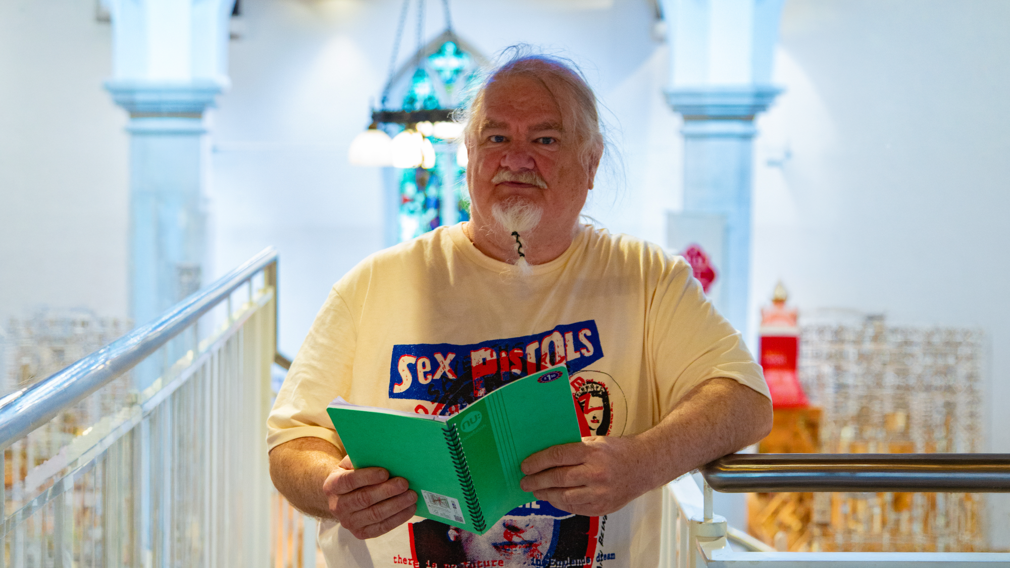 Trivallis Housing Landlord Wales An older man with white hair and a short beard stands indoors holding an open green notebook. He is wearing a Sex Pistols T-shirt. Decorative architecture and a stained glass window are visible in the background.