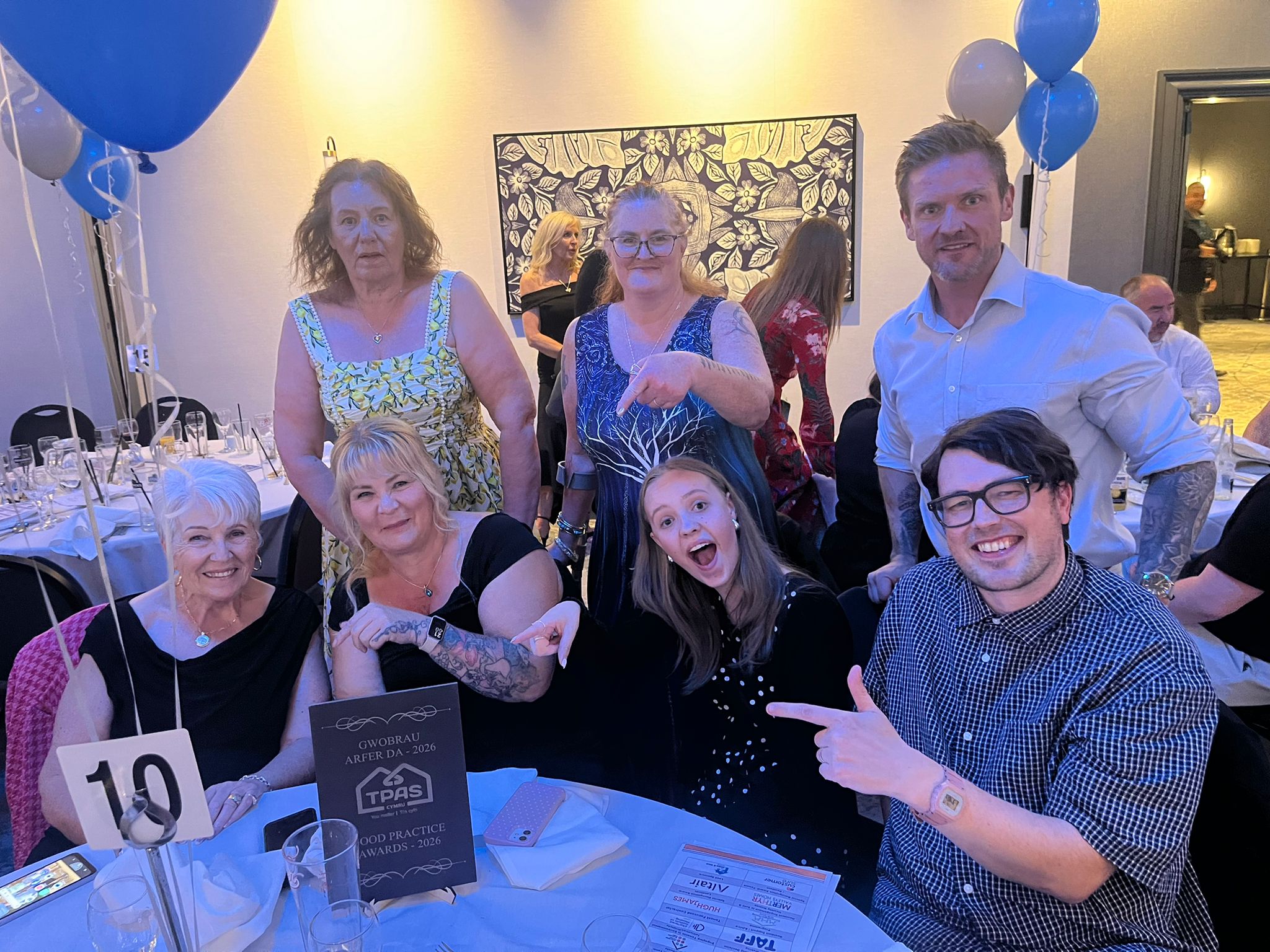 Trivallis Housing Landlord Wales Seven adults pose and smile around a dining table at a formal event. The table holds glasses, plates, a plaque, and a “10” number sign. Blue balloons are visible and other guests are present in the background.