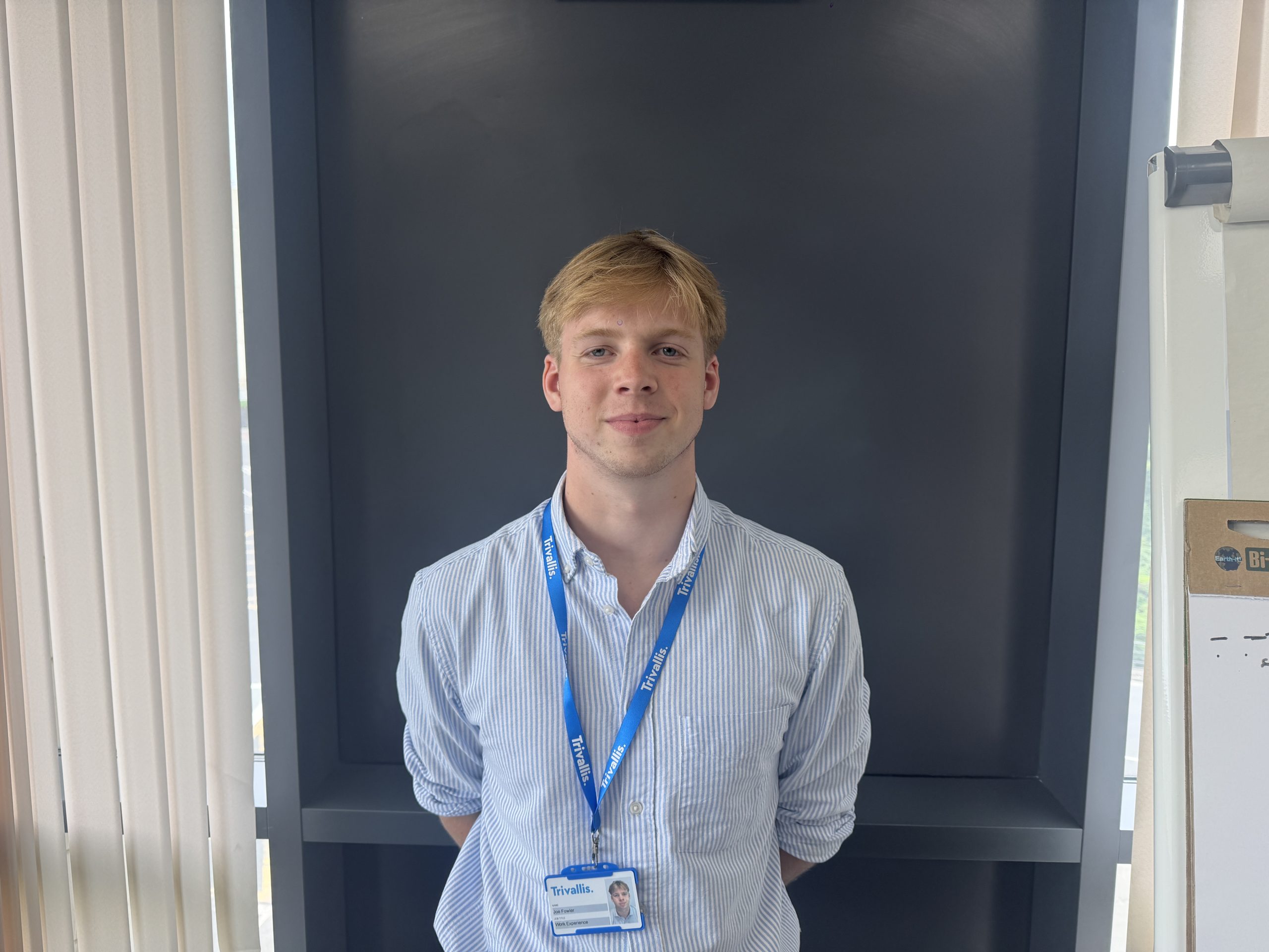 Trivallis Housing Landlord Wales A young man with light hair wearing a blue striped shirt and a blue lanyard stands in front of a dark gray wall, with his hands behind his back. The room has vertical blinds and part of a whiteboard is visible on the right.