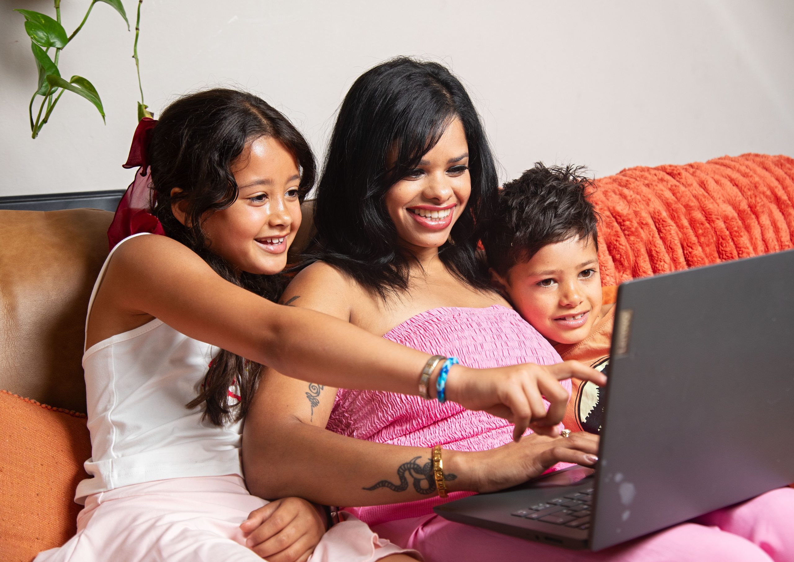 Trivallis Housing Landlord Wales A woman and two children sit closely together on a couch, smiling and looking at a laptop screen. One child is pointing at the laptop. They appear to be engaged with what is on the screen.