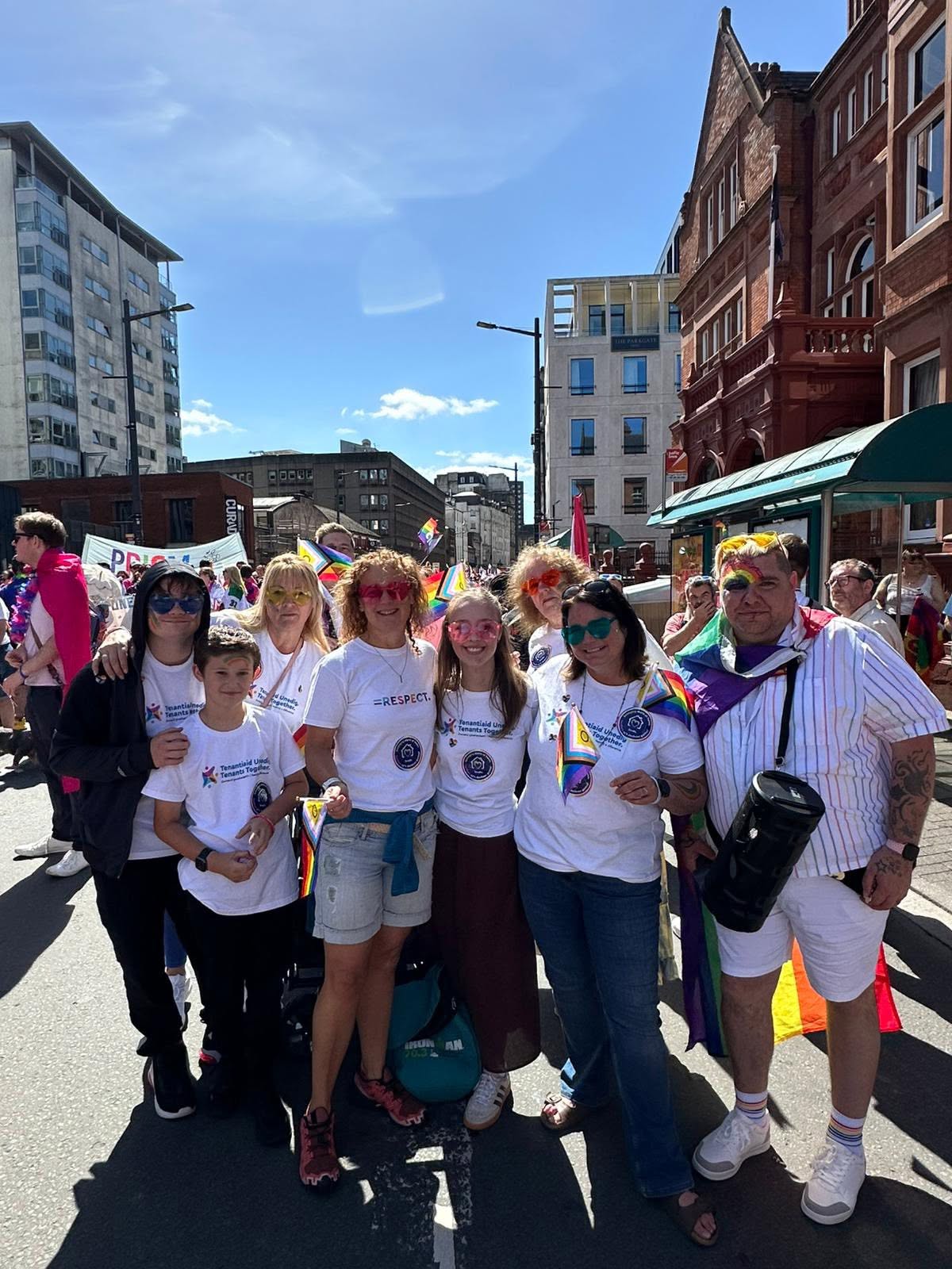 Trivallis Housing Landlord Wales A group of people wearing matching white T-shirts and holding rainbow flags pose together on a sunny street during a parade. Buildings and other participants are visible in the background.