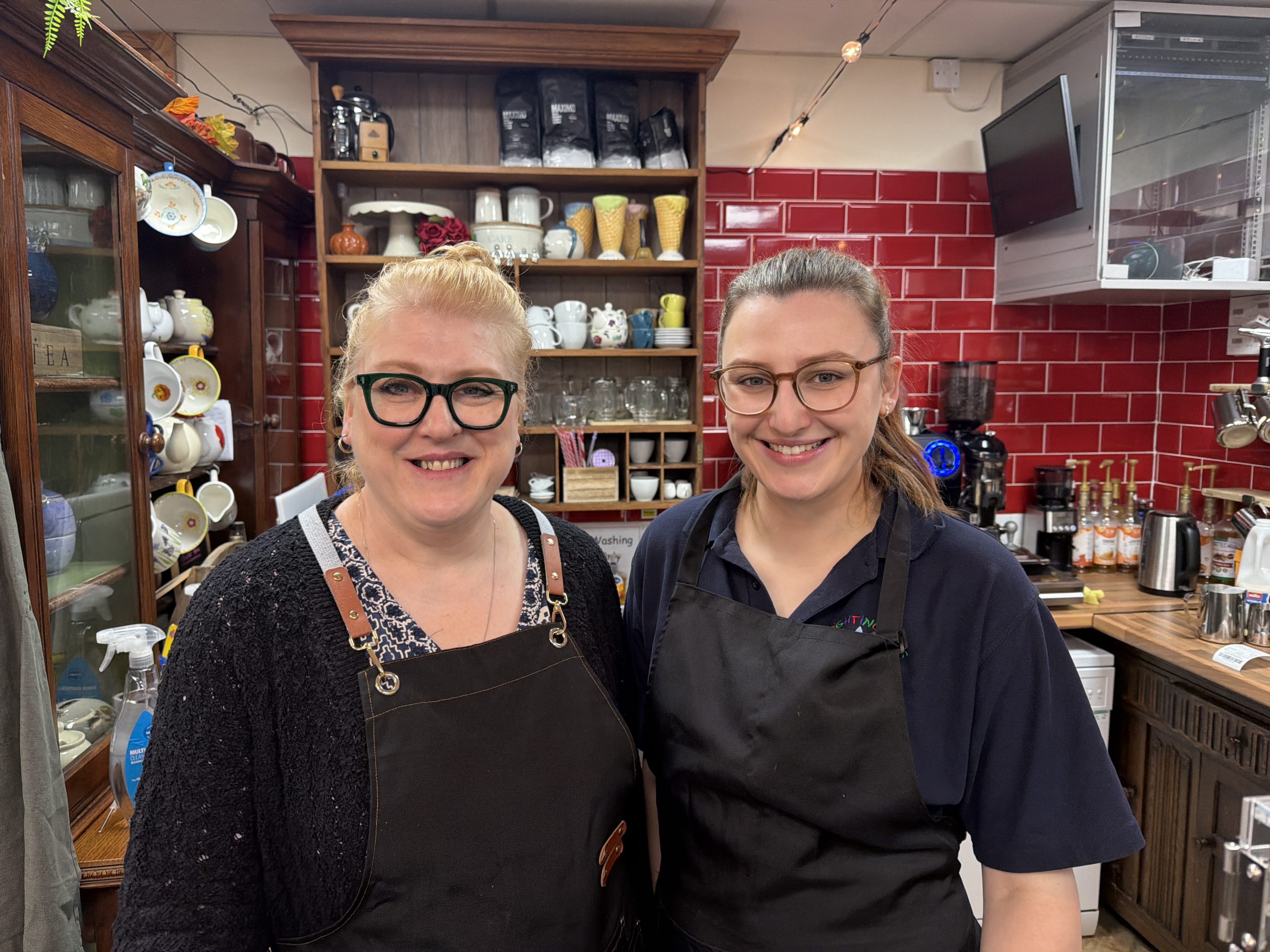 Trivallis Housing Landlord Wales Two people wearing black aprons stand side by side in a café with red tiled walls, glassware, cups, and coffee equipment visible behind them. Both are smiling and looking at the camera.