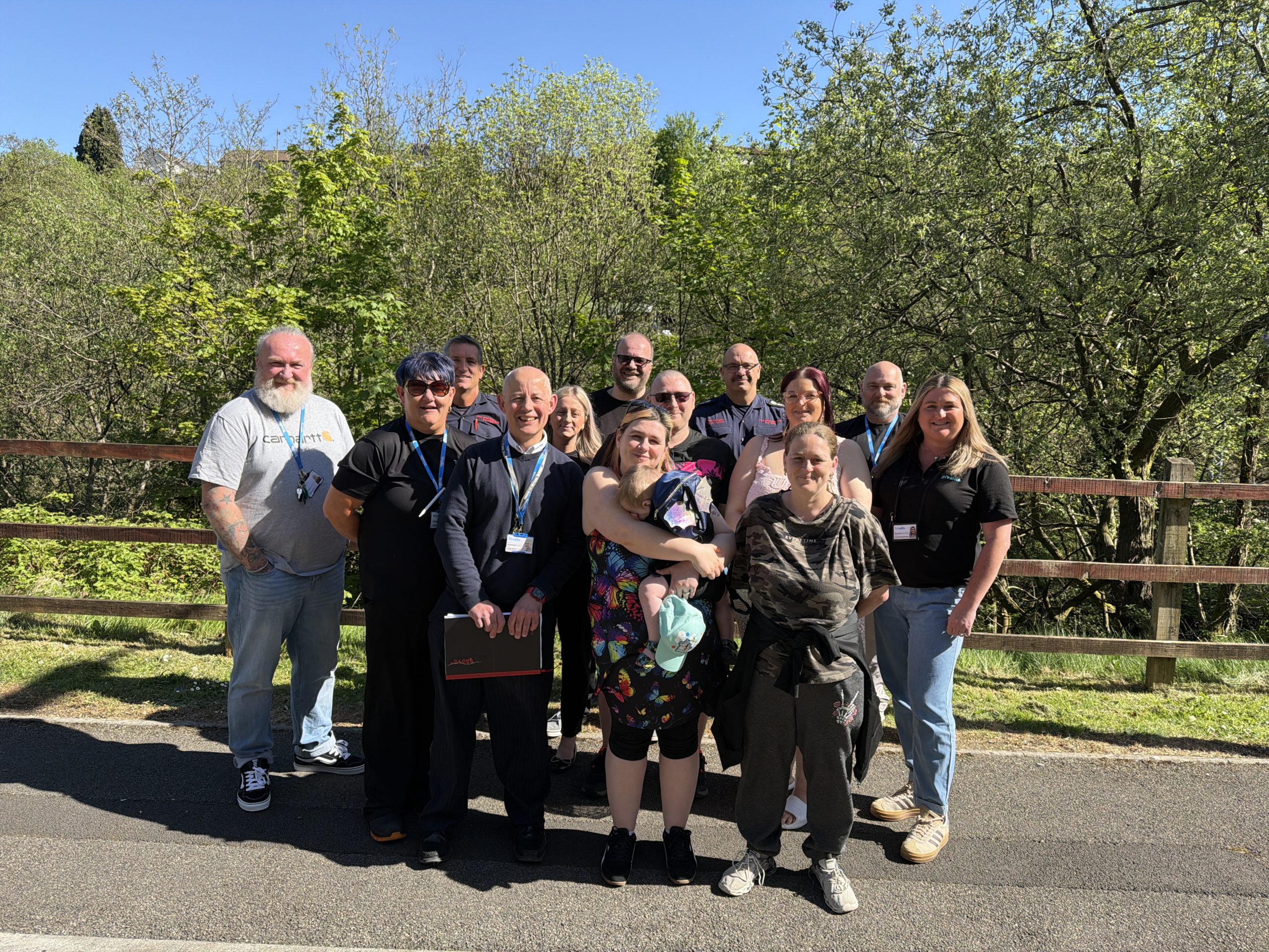 Trivallis Housing Landlord Wales A group of fifteen people stand together outdoors on a sunny day, posing for a photo in front of a wooden fence with green trees and foliage in the background.