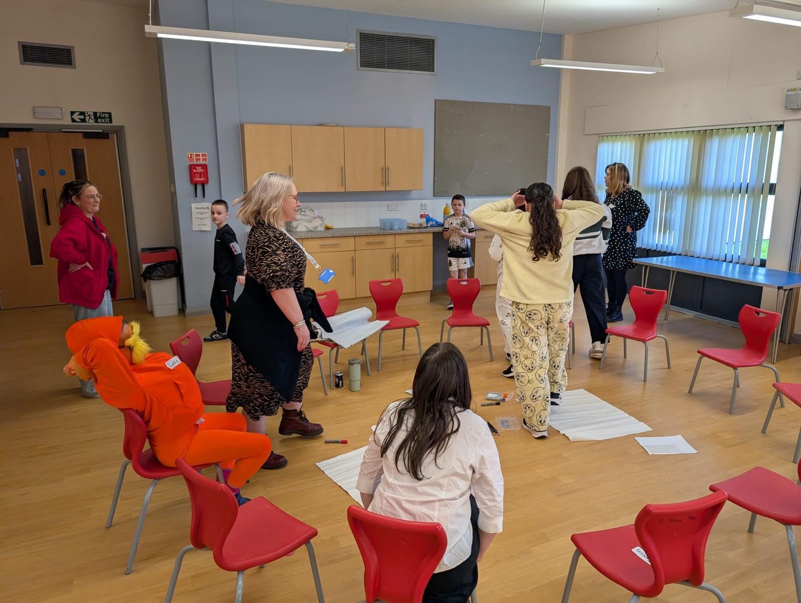 Trivallis Housing Landlord Wales A group of people, mostly girls, stand and sit on red chairs arranged in a circle in a room with wooden floors and light blue walls. Some hold papers, and one person is dressed in an orange costume.